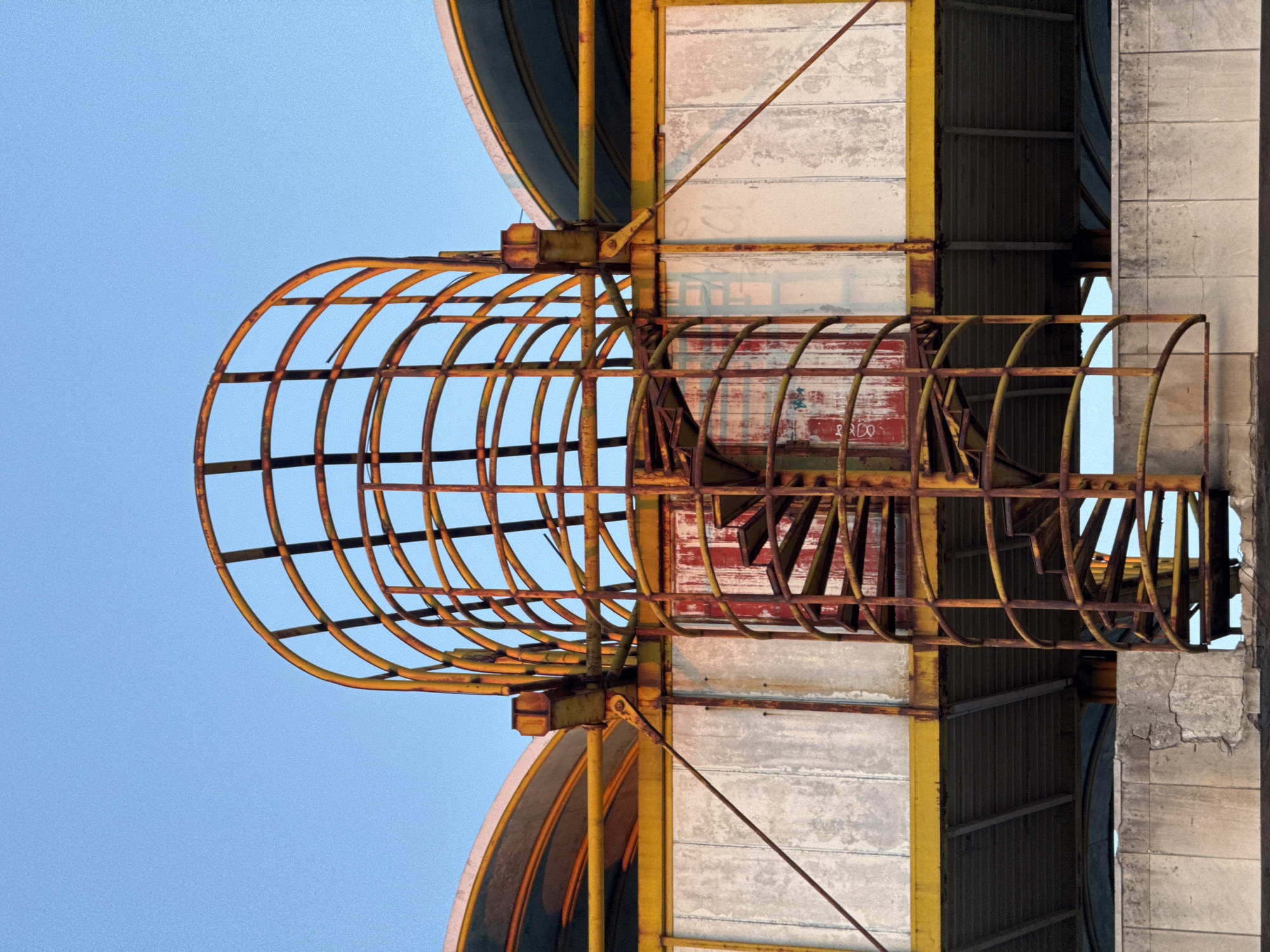 A low-angle, vertically-oriented shot reveals a rusting metal structure against a pale blue sky. The structure is composed of cylindrical cages made of yellowed and rusted bars, attached to a building. The building is constructed of concrete panels with yellow framing. One cylindrical cage is prominently visible on the left, extending outwards and upwards towards the sky. Behind it and slightly to the right, a similar structure is partially obscured. The building itself shows signs of aging and weathering. The overall impression is one of industrial decay and abandonment. This specific structure is part of the Tabaena Brewery in the city of Plovdiv, Bulgaria.