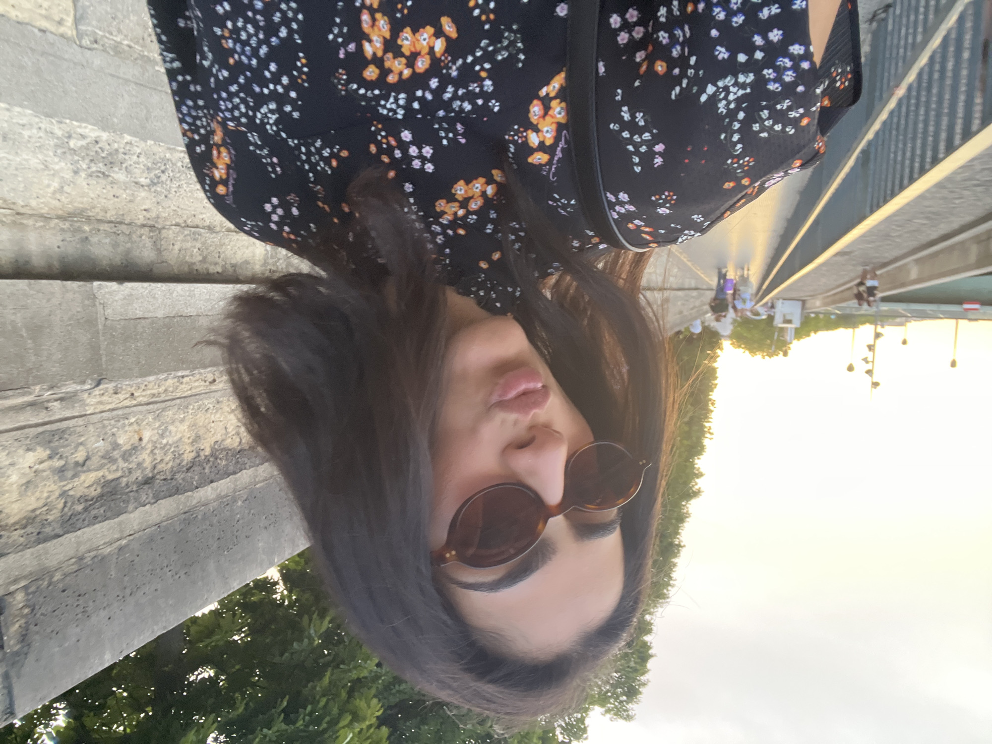 A woman with dark hair and oval sunglasses is taking a selfie while walking outdoors. She is wearing a black dress with a floral pattern. In the background, there is a stone wall, a tree, a walkway with people, a railing, and a body of water. The sky is bright, suggesting it is daytime or sunset. The overall scene appears to be in an urban environment, possibly along a river in Paris, France, given the architecture and pedestrian walkway.