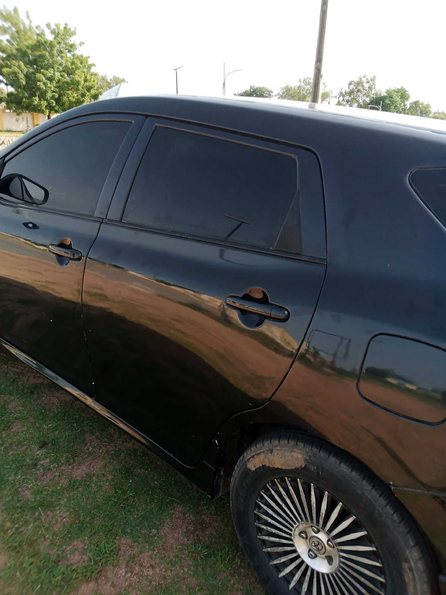 The image shows the side of a black Toyota Corolla parked on a grassy area, possibly in Jalingo, Nigeria. The car has tinted windows and a shiny black exterior that reflects the surroundings. The wheel in the foreground is visible, with a silver rim and a tire that appears to have some dirt on it. In the background, there are trees and a bright sky, suggesting an outdoor setting. The overall impression is of a car parked in a somewhat casual environment, with the reflection on the car's surface indicating an open area.