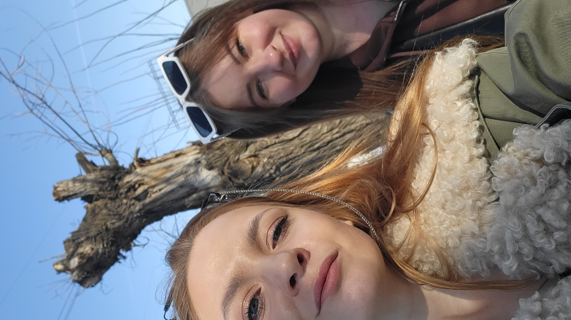 Two women are in a selfie together. The woman on the bottom has red hair and is wearing small sunglasses. The woman on top has brown hair and is wearing white-framed sunglasses. Behind them is a tree and a clear blue sky. The location may be Chaikovsky, Russia.