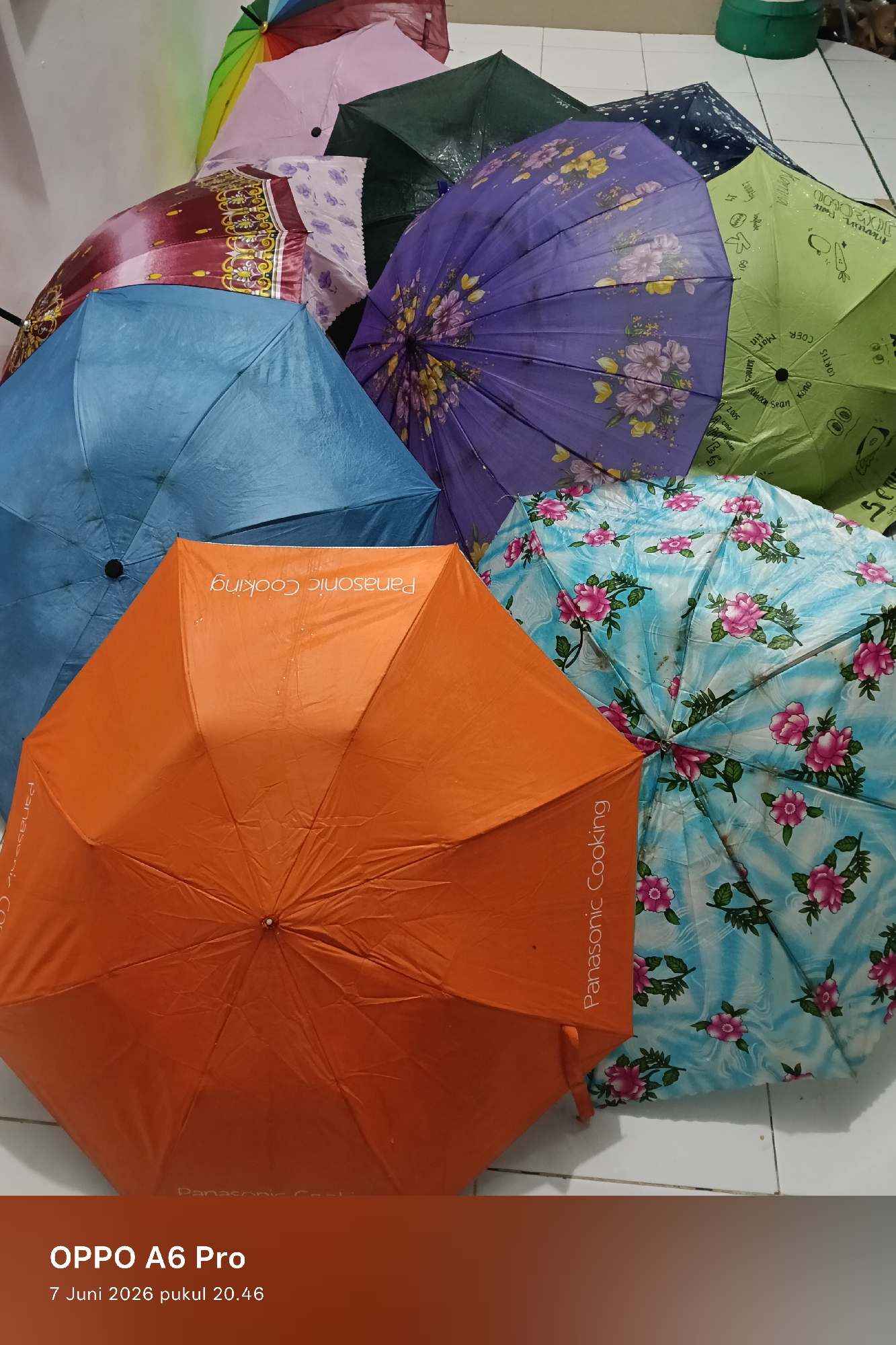 The image depicts a collection of multiple open umbrellas laid out on a light-colored tiled floor, leaning against a plain white wall, taken in what is understood to be the city of Lhokseumawe, Indonesia. No individuals are present in the scene.
The umbrellas display a variety of colors and patterns:
* A partially visible multi-colored, possibly rainbow-striped, umbrella is at the top left.
* Below it, a solid pink umbrella is seen.
* A maroon umbrella with intricate gold, yellow, and white floral or abstract patterns is spread open.
* A dark green, solid-colored umbrella is visible.
* A dark blue umbrella with white polka dots is partially obscured.
* A prominent purple umbrella features large, detailed floral patterns in shades of pink, yellow, and white.
* A large light blue umbrella is visible, showing signs of wear and possibly dirt.
* A large orange umbrella, centrally positioned, has "Panasonic Cooking" printed in white text; the text appears inverted on one section due to the umbrella's folds.
* A light green umbrella, distinctively patterned with various whimsical black line drawings including text such as "COOK," "MOR," "JAWAB," a carrot, and an egg.
* A light blue umbrella with vibrant pink floral designs and green leaves covers the lower right portion of the frame, also appearing to have water droplets or smudges on its surface.
The umbrellas are arranged in an overlapping manner, suggesting they are either being dried or stored. The tiled floor is a light, possibly white or off-white, hue. In the background to the right, a dark green circular container or bucket is partially visible on the floor, next to some indistinct brown objects. The lighting suggests an indoor setting, consistent with the captured time.
The image metadata indicates it was captured by an "OPPO A6 Pro" on "7 Juni 2026 pukul 20.46" (June 7, 2026, 8:46 PM).