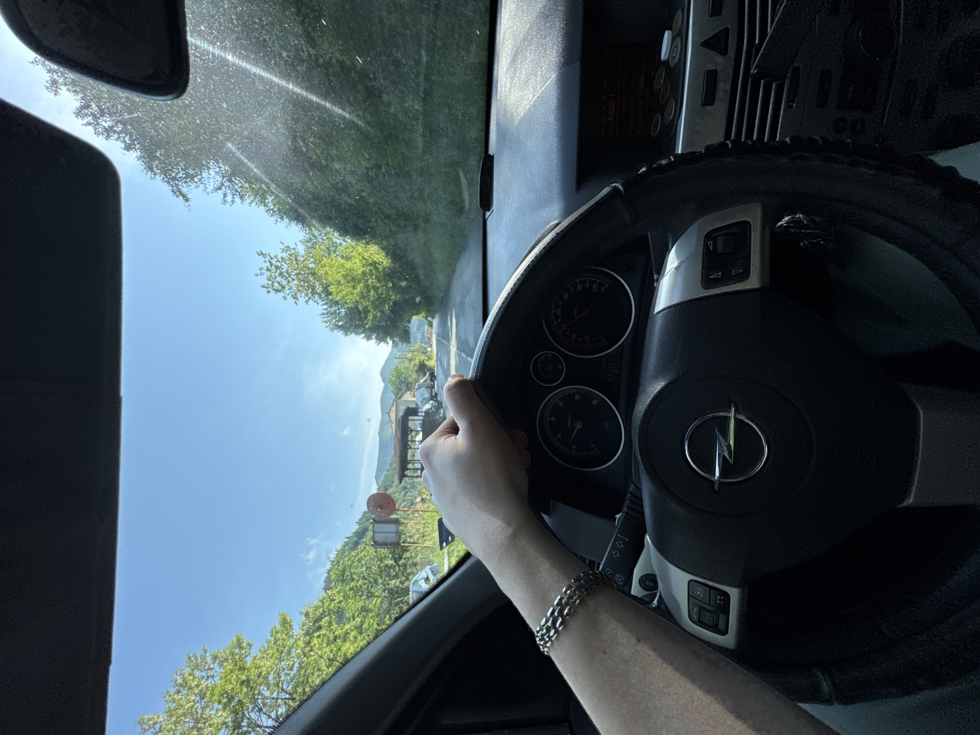 A vertically oriented image captures a view from inside a car, likely from the driver's perspective, though the scene outside appears rotated with the horizon running vertically along the left. A human right arm and hand are visible in the foreground, gripping the black steering wheel. The arm wears a silver-colored chain bracelet.
The steering wheel, featuring a silver Opel logo (a lightning bolt in a circle), is prominent in the mid-right of the frame. Parts of the car's dashboard, including the instrument cluster with a speedometer (showing markings like 100, 140, 180, 220 km/h), air vents, and a radio/display unit with the text "RADIO 1" are discernible. Two sun visors are partially visible at the top left.
Outside the car, through the windshield, a bright and sunny day is depicted with a clear blue sky. Lush green trees line what appears to be a road, and distant mountains or hills are visible. A red and white circular sign stands on the left side of the road, and some buildings or structures are seen in the background.
The activity taking place is driving a car through a scenic, rural or mountainous area. The bright lighting suggests it is daytime. The location is Taran, Bulgaria.