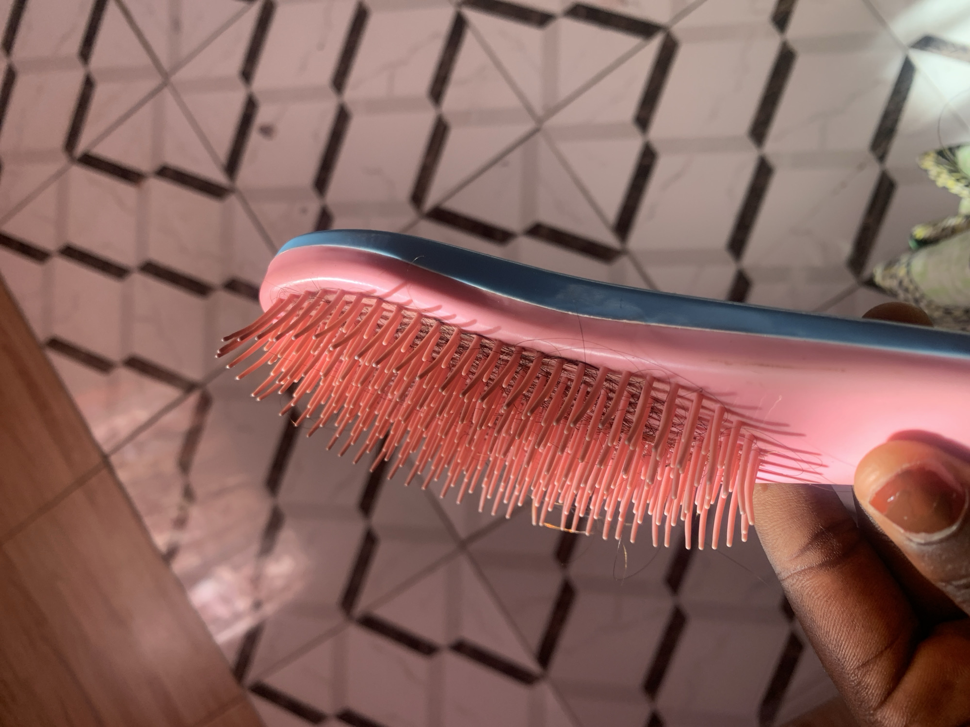 In Jauro Yinu, Nigeria, a close-up shot focuses on a pink and blue hairbrush held by a hand with dark skin. The brush's bristles are pink, and several strands of dark hair are visible on them. The background features a tiled floor with a geometric pattern of light and dark squares. Sunlight casts shadows across the tiles.