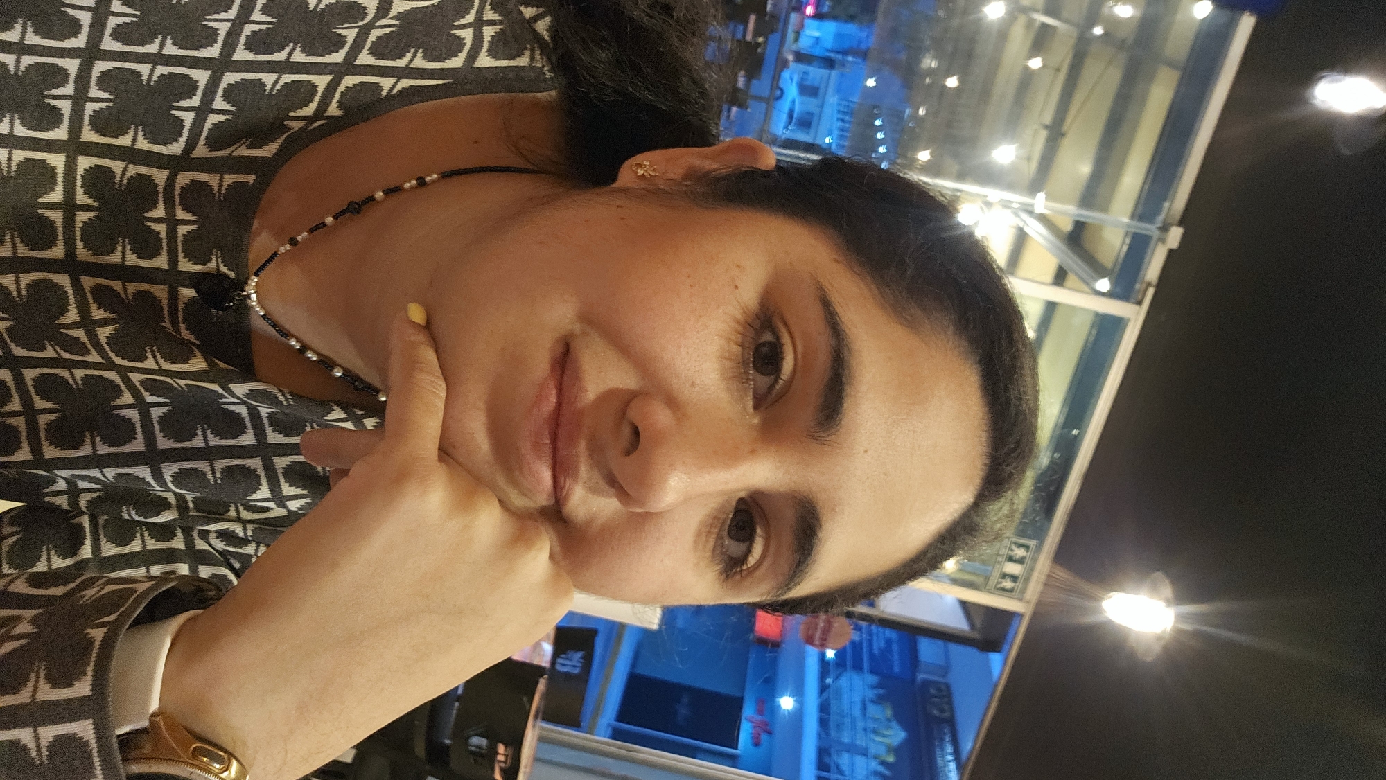 A woman with dark hair and medium skin tone is looking at the camera. She is wearing a patterned gray and white shirt, a gold watch, and a beaded necklace. Her hand is touching her face. She appears to be indoors at night. Visible through the window behind her is a city scene, likely Puebla, Mexico, with illuminated buildings and lights. The location appears to be inside a restaurant at night.