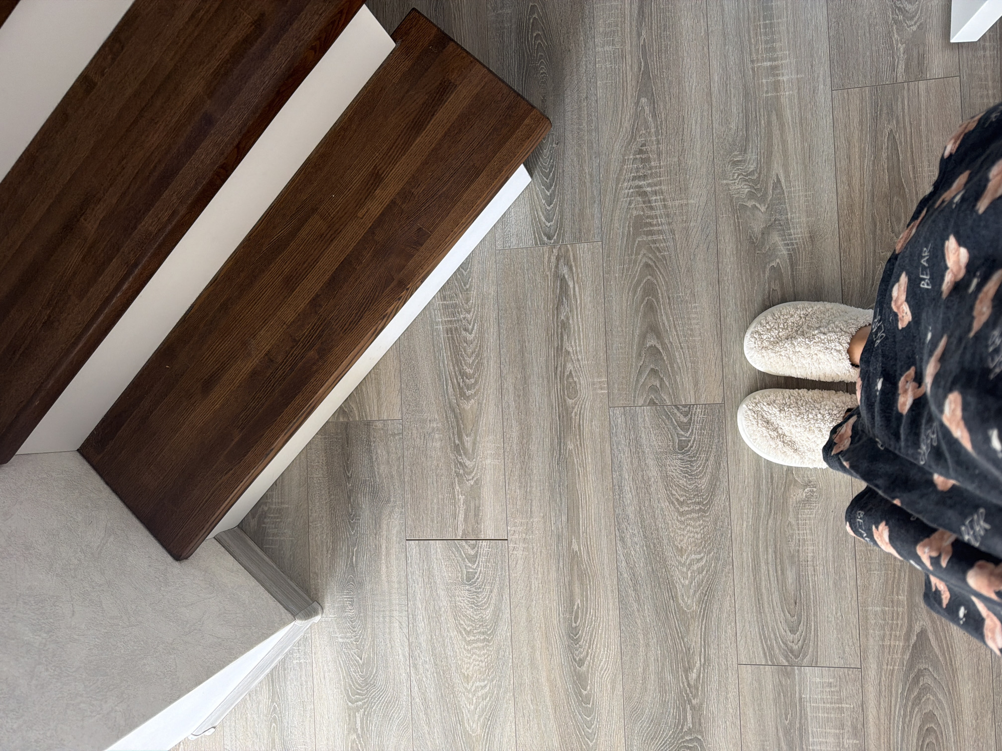 A top-down, high-angle shot captures a pair of feet wearing fluffy white slippers, standing on a light-colored, wood-grain laminate floor. The feet are clad in dark pajamas adorned with a repeating pattern of cartoonish bears and the word "BEAR". In the background, a wooden staircase with dark brown treads and white risers ascends. The wall adjacent to the staircase has a textured, light gray wallpaper. The scene appears to be an indoor residential setting, possibly in Malinovka, Russia. There are no other people or discernable activities. The lighting is bright and even, suggesting natural light or well-lit interior conditions.