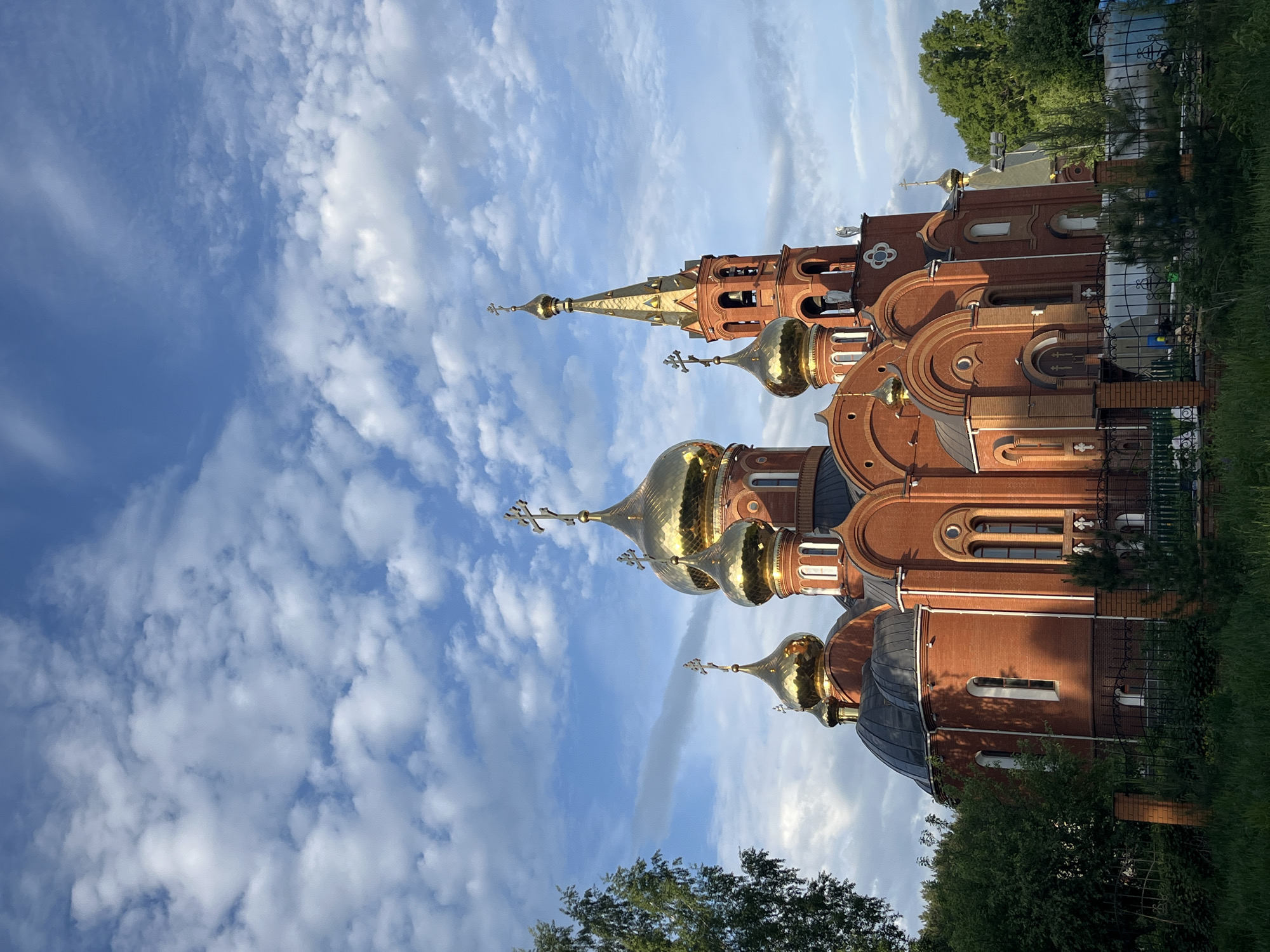The image depicts the Holy New Martyrs and Confessors of Russia Temple, an Orthodox church located in Chaikovsky, Perm Krai, Russia. The temple features a red brick facade with golden domes and crosses on top. The sky is blue with scattered white clouds. Trees and a fence are visible around the temple grounds.