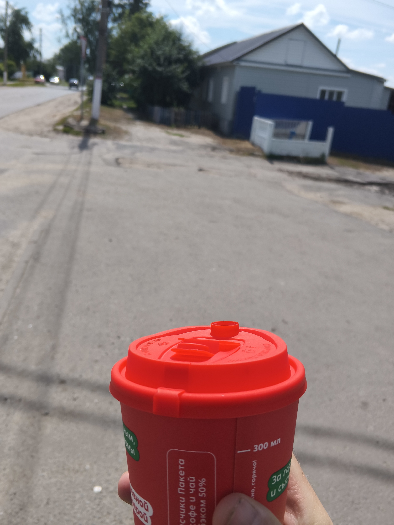 The image shows a red coffee cup with a red lid held by a person's hand. Text on the cup is visible, including "300 мл" and some Russian phrases. The background suggests an outdoor urban environment. In the distance, there is a road with tire tracks, a sidewalk, a utility pole, trees, and a building with a blue fence. The scene appears to be a street in Mokshan, Russia, based on the presence of Russian text on the cup and the characteristic architecture of the building. The sky is partly cloudy.