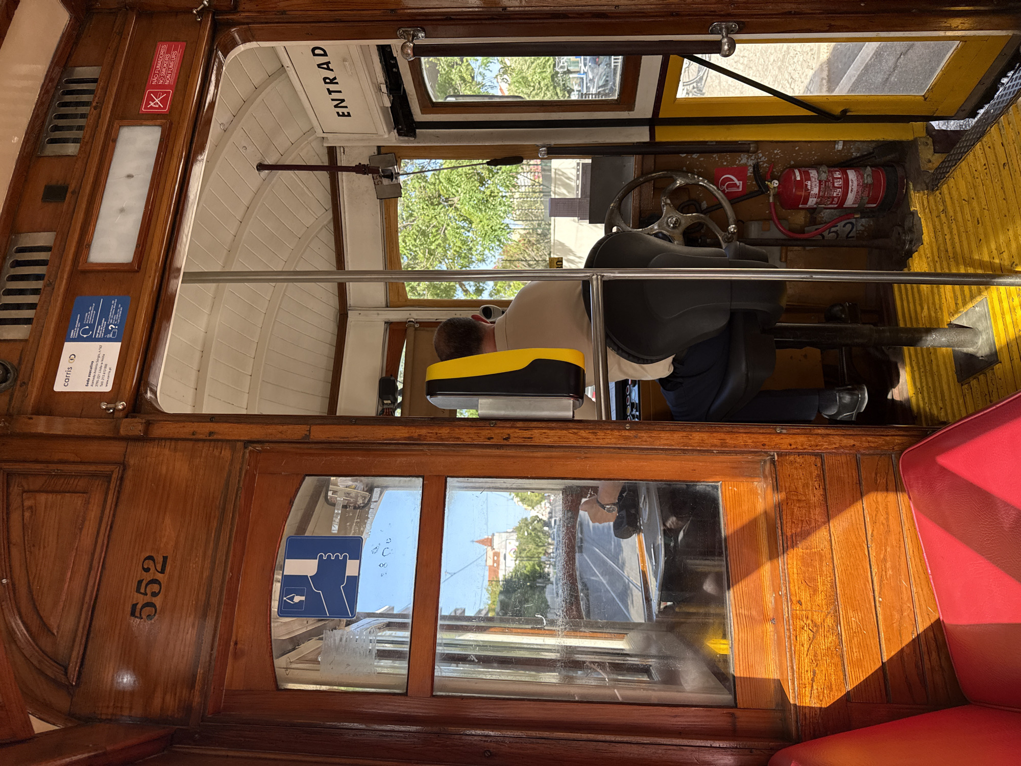 The image shows the interior of a vintage tram, specifically Tram 552, in Lisbon, Portugal. The tram's interior is characterized by rich, polished wooden paneling and red upholstered seating. The driver's area is visible, with the driver seated at the controls, wearing a light-colored shirt and dark trousers. A fire extinguisher is mounted on the wall. Through the windows, a street scene in Lisbon can be observed, with buildings, trees, and other trams. The lighting suggests it is daytime, with sunlight casting shadows on the wooden interior. The overall impression is one of a well-maintained, historic public transport vehicle.