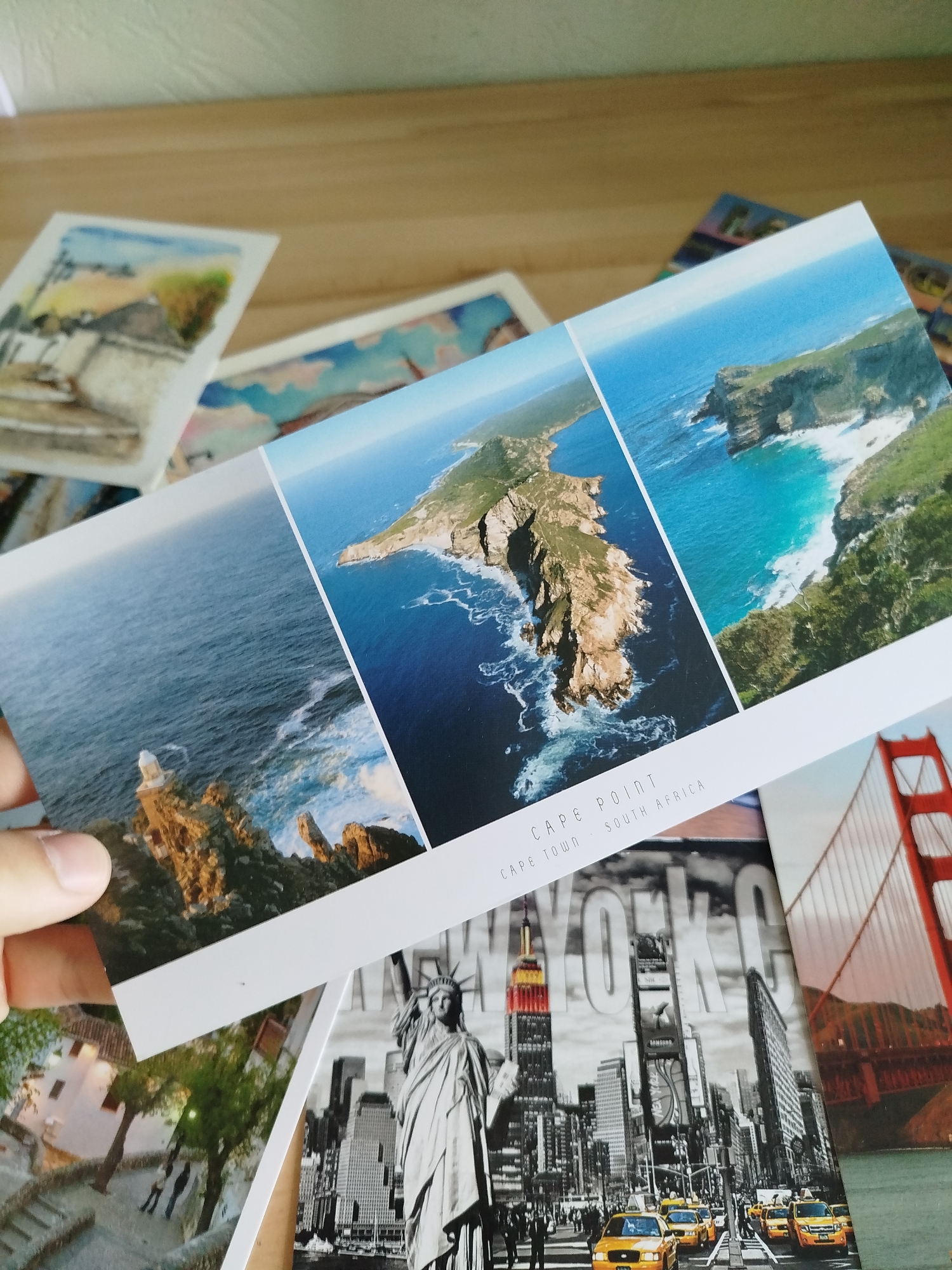 A person's hand is holding a panoramic postcard that displays three images of a coastal landscape. The postcard is placed over other postcards, one of which shows a black and white cityscape with the Statue of Liberty and yellow taxis, likely representing New York City. Another postcard partially visible features the red suspension of a bridge, possibly the Golden Gate Bridge. The dominant postcard features rugged cliffs meeting the ocean, with a lighthouse on a rocky outcrop. Text on the postcard reads "CAPE POINT CAPE TOWN SOUTH AFRICA". The scene is set on a wooden surface. The person's hand is positioned to hold the postcard, obscuring a portion of the bottom left corner. There are no identifiable elements within the image that suggest the location is Varna, Bulgaria.