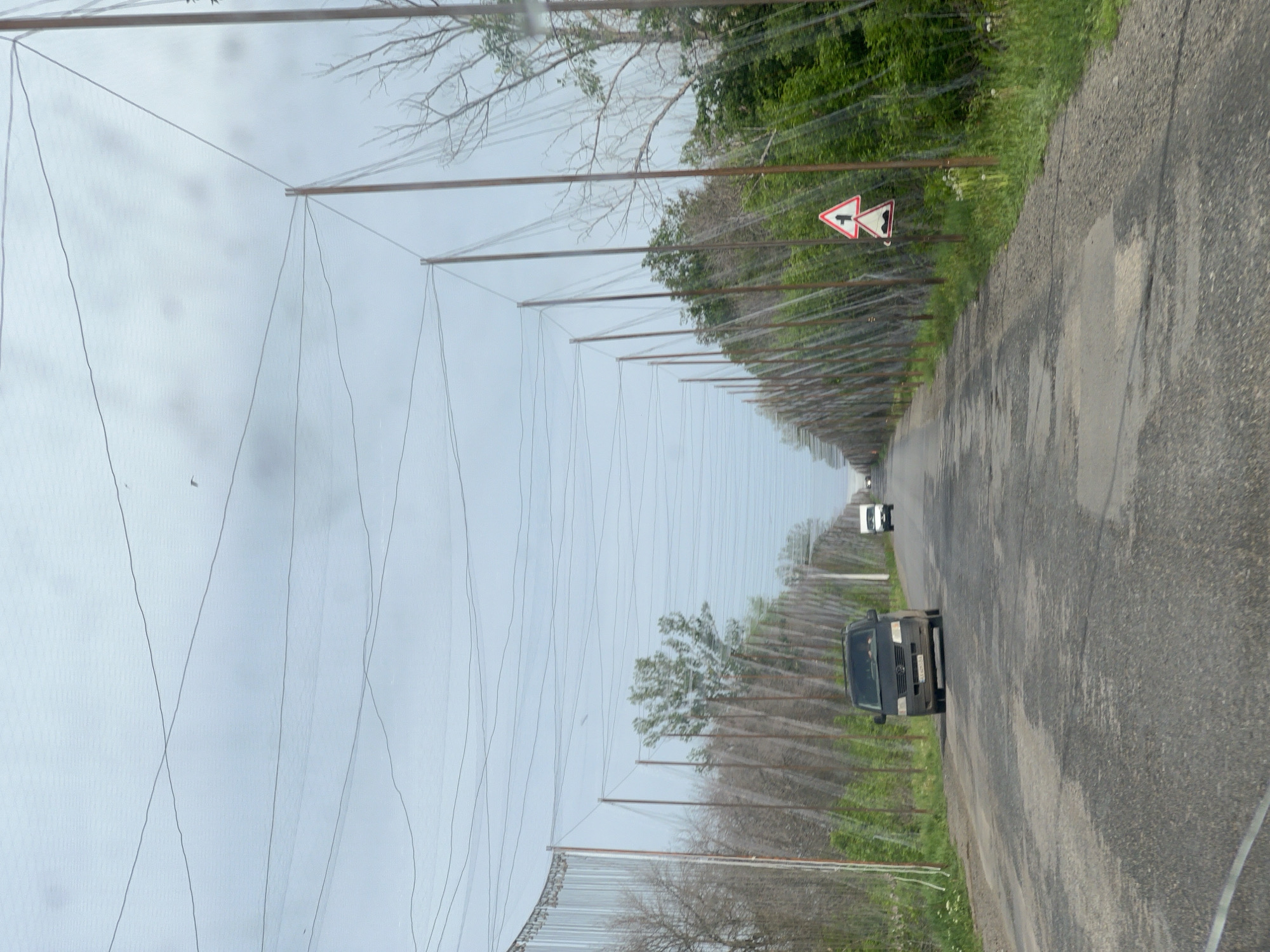 The image shows a rural road in Derhachi, Ukraine. A long row of poles covered with netting stretches along both sides of the road, creating a tunnel-like effect. A black van is parked on the right side of the road, and another vehicle is visible in the distance. A road sign indicating a side road junction is present on the right side of the road. The road surface is cracked and uneven. Vegetation lines the edges of the road. The sky is overcast.