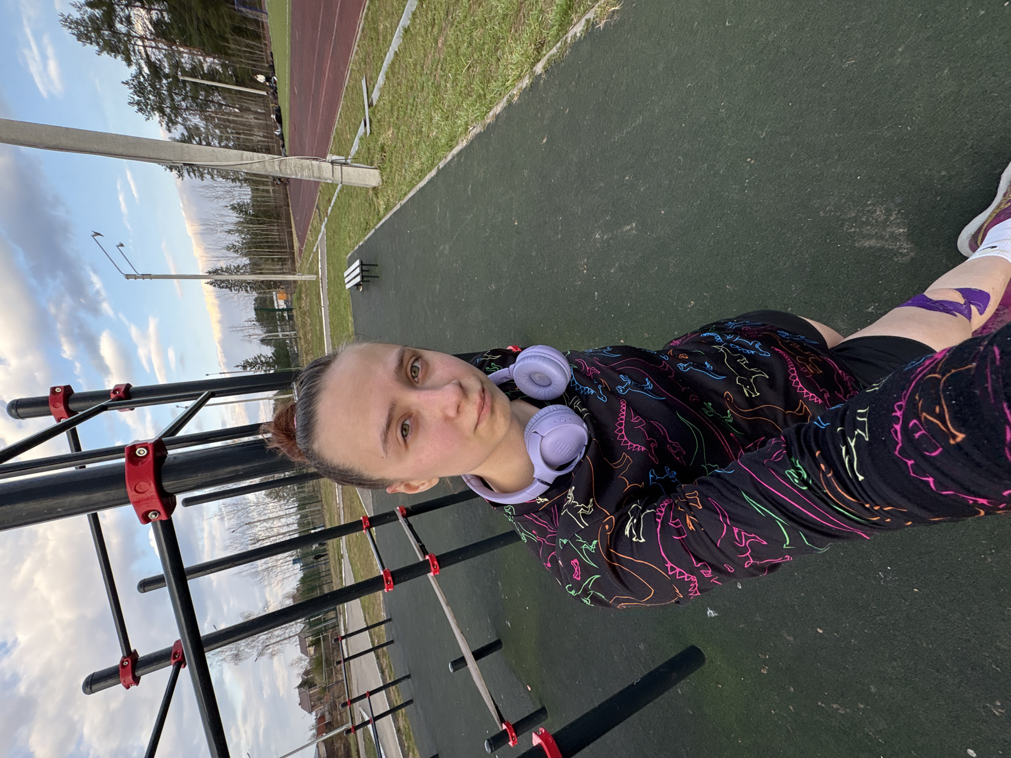 A woman stands on a dark green surface at an outdoor fitness area, possibly in Прикамский, Russia. She is wearing a black long-sleeved shirt with colorful dinosaur patterns and has purple headphones around her neck. Her hair is pulled back. A piece of purple athletic tape is visible on her right knee. In the background, there's exercise equipment including monkey bars with red fittings. Beyond that, a running track and trees are visible. The sky is light with clouds.