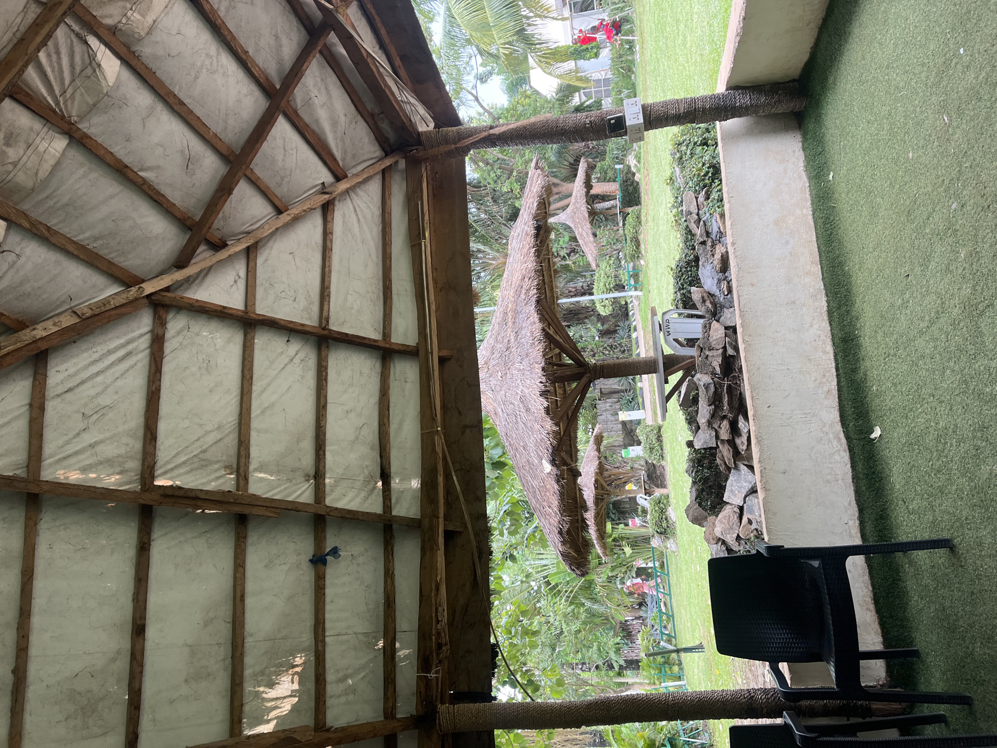 The image displays an outdoor recreational area in Galadimawa Village, Nigeria, observed from within a covered pavilion.
The immediate foreground features a rustic ceiling constructed from rough-hewn wooden beams forming a grid, covered by a light-colored, possibly white or gray, tarpaulin or fabric material that shows signs of weathering, creasing, and minor staining. A small blue plastic object or bag is suspended from a central beam. Two thick, cylindrical support pillars are visible, wrapped in a coarse, natural fiber material. The pillar on the right has a white electrical power strip with three visible outlets attached to it. The floor of this area is covered with artificial green turf. A black plastic chair with armrests occupies the left side of the turf, with a portion of another identical chair visible to its left. A low, light-colored concrete or stucco wall separates the foreground seating area from the lawn beyond. This wall features a small rock arrangement and some greenery on its surface, positioned between the wall and the main lawn.
The middle ground consists of a well-maintained green lawn. Several structures with conical thatched roofs, resembling umbrellas or small huts, are distributed across the lawn. A prominent thatched umbrella with a visible wooden base/table is centrally located. To its left, a smaller thatched umbrella is present, with a white chair or table base underneath. Another thatched structure is visible further back on the right.
Multiple individuals are present on the lawn. On the left side, a person in a light-colored, possibly red or pink, long-sleeved outfit is walking from left to right. On the far right, near a tall palm tree, two figures are visible; one wears a red top, standing near a red metallic structure. Various trees, including palm trees and dense green foliage, are scattered throughout the lawn area. A white sign with the letters "WWP" is partially visible near the base of a table to the left of the central thatched umbrella.
The background shows further dense green vegetation, with taller trees and bushes, and a faint glimpse of a white building through the foliage on the far right. The overall lighting suggests daylight conditions, with some dappled light visible on the ceiling.
