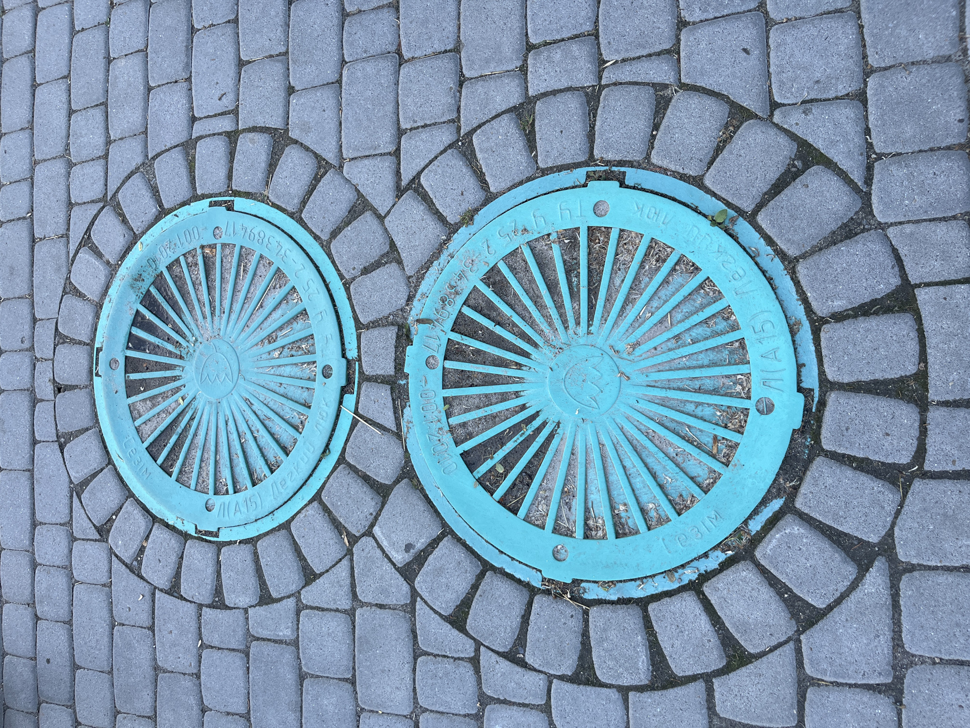 The image shows a close-up, overhead view of two turquoise manhole covers set into a cobblestone pathway. The manhole covers are round and feature a radial spoke pattern. Text and numbers are visible on the rims of both covers, although they are difficult to read clearly in the original image. One cover has "LE31M" and "(A15)" visible. The other cover has "0012010" and "2-34389417" visible. The cobblestones are a mix of gray and dark gray rectangular and square shapes. Some dried leaves and dirt are present on the ground around and between the manhole covers. The image appears to have been taken outdoors, likely on a street or pedestrian area.