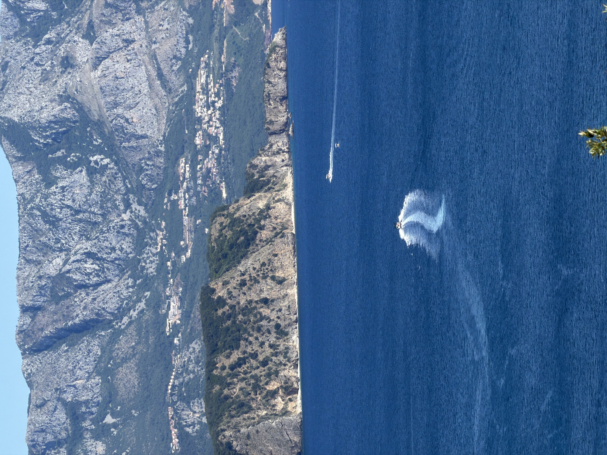 The image depicts a scenic view of the Adriatic Sea near Dubraljevina, Montenegro. In the foreground, the deep blue water stretches towards the horizon. Two white speedboats are visible, leaving white wakes behind them as they move across the water. In the mid-ground, a rugged, tree-covered cliff face juts out into the sea. Behind the cliff, a town with white-roofed buildings is nestled on the slopes of a large, imposing mountain range. The mountains are rocky and sparsely vegetated, with a clear blue sky overhead. The overall scene conveys a sense of natural beauty and tranquility.
