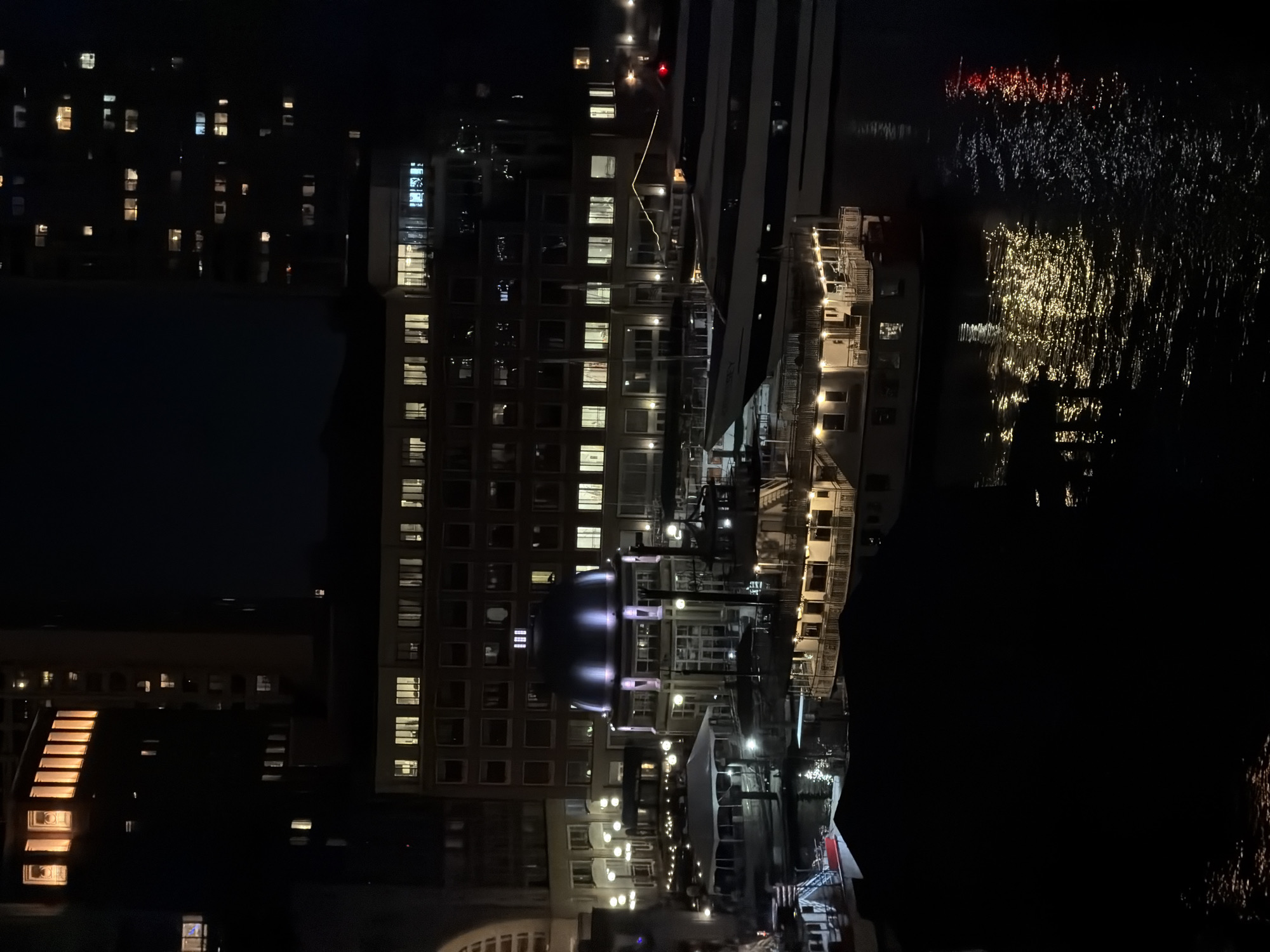The image is a night view of Boston, United States. The primary subject is a cluster of buildings, predominantly high-rise structures illuminated with numerous window lights. A distinctive building with a rounded dome-like structure stands out amidst the angular skyscrapers. The dark expanse possibly indicates a body of water, likely the harbor, with reflections of distant lights creating a shimmering effect. There is minimal presence of visible people in the scene. The environment is characterized by the urban landscape at night, with varying levels of illumination and shadow play.