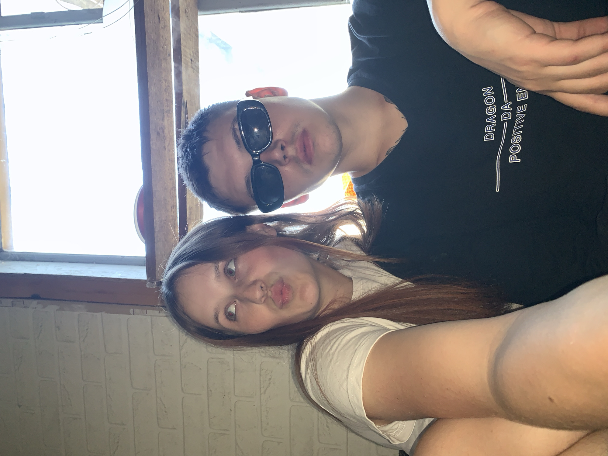 Two people are posing for a selfie in an indoor environment. The person on the left is a young woman with long, light brown hair wearing a white t-shirt and has a playful expression on her face, with pursed lips. The other person on the right is wearing sunglasses and a black t-shirt with white writing on it. A window with a wooden frame can be seen in the background, with a white brick wall partially visible near the bottom of the image. The exact location in Фоки, Russia, cannot be determined from the given image.