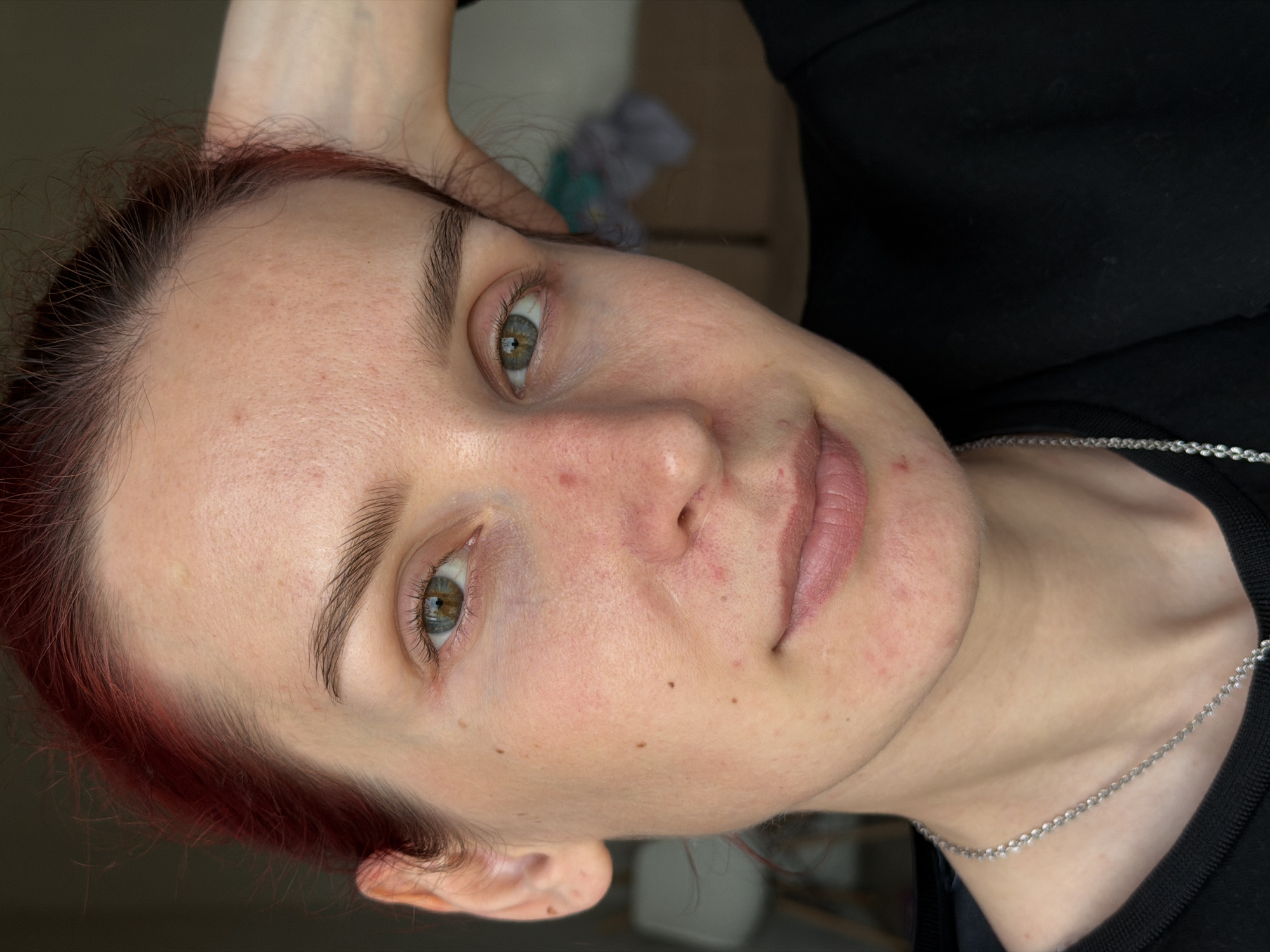 A close-up portrait of a woman with fair skin and red hair is presented at an angle. Her eyes are hazel-green and she has dark eyebrows. There are some blemishes on her face. She is wearing a black shirt and a silver chain necklace. Part of her hand is visible near her head. The background is slightly blurred and shows indistinct objects and a wall. There is no definitive evidence in the image to confirm that it was taken in Chaikovsky, Russia.