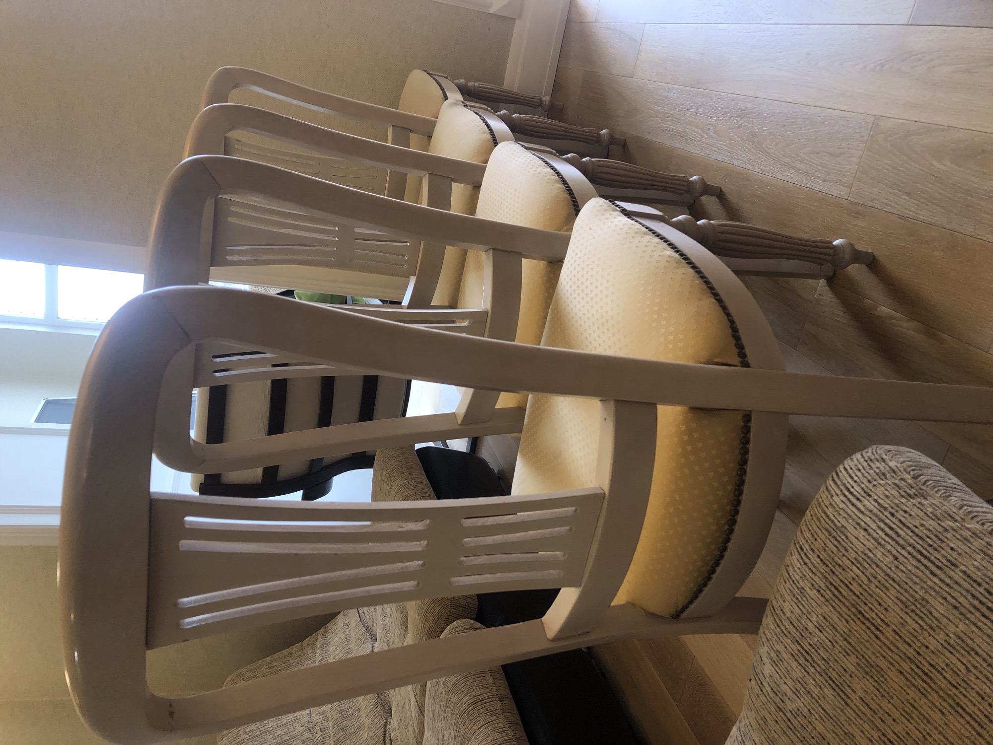 The image shows a row of three light-colored wooden chairs with yellow upholstered backs, stacked one behind the other. The chairs have curved backs with horizontal slats. The environment appears to be indoors, with a light-colored wall and a wooden floor in the background. There is a window visible in the distance, providing natural light. No people are visible in the frame. There is nothing to indicate that this scene is in the city of Chaikovsky, Russia.