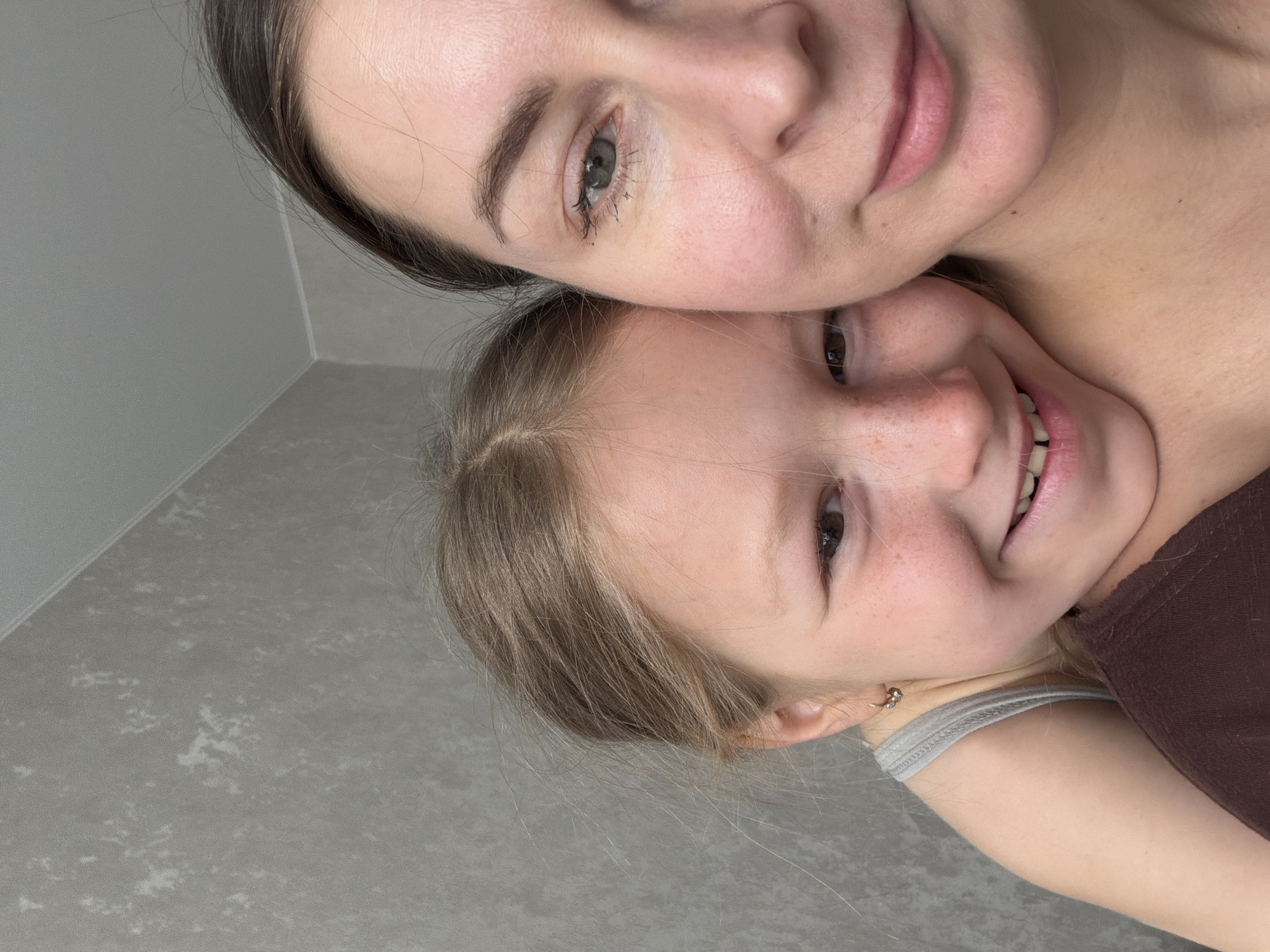 Two individuals, likely a mother and daughter, are posing closely for a selfie indoors. The adult has dark hair, fair skin, and is wearing no visible jewelry. The child has blonde hair, fair skin with freckles, and is wearing a light-colored tank top under a brown garment. The background consists of a plain wall and floor. The location is potentially Dubovaya, Russia.
