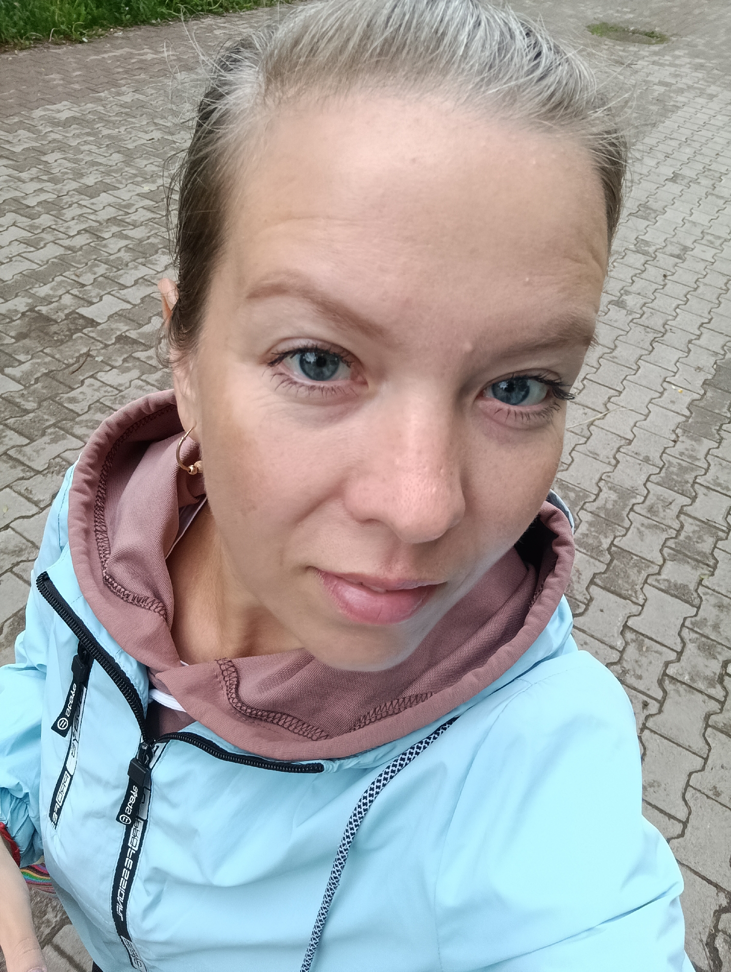 The image depicts an adult female positioned outdoors in the city of Chaikovsky, Russia. She has fair skin, blue eyes, and light brown hair with visible gray strands, pulled back from her face. Her expression is neutral, with her gaze directed at the camera and her lips slightly parted. A small, gold-colored hoop earring is visible in her right ear. She is wearing a light blue hooded jacket, which features black zippers and striped black and white webbing with the text "ЯЕЛЕВ€" (reversed) along the zipper line, layered over a pinkish-brown hooded sweatshirt. The background shows a paved surface of gray rectangular interlocking pavers, which appears damp, and a strip of green grass and dirt further back.