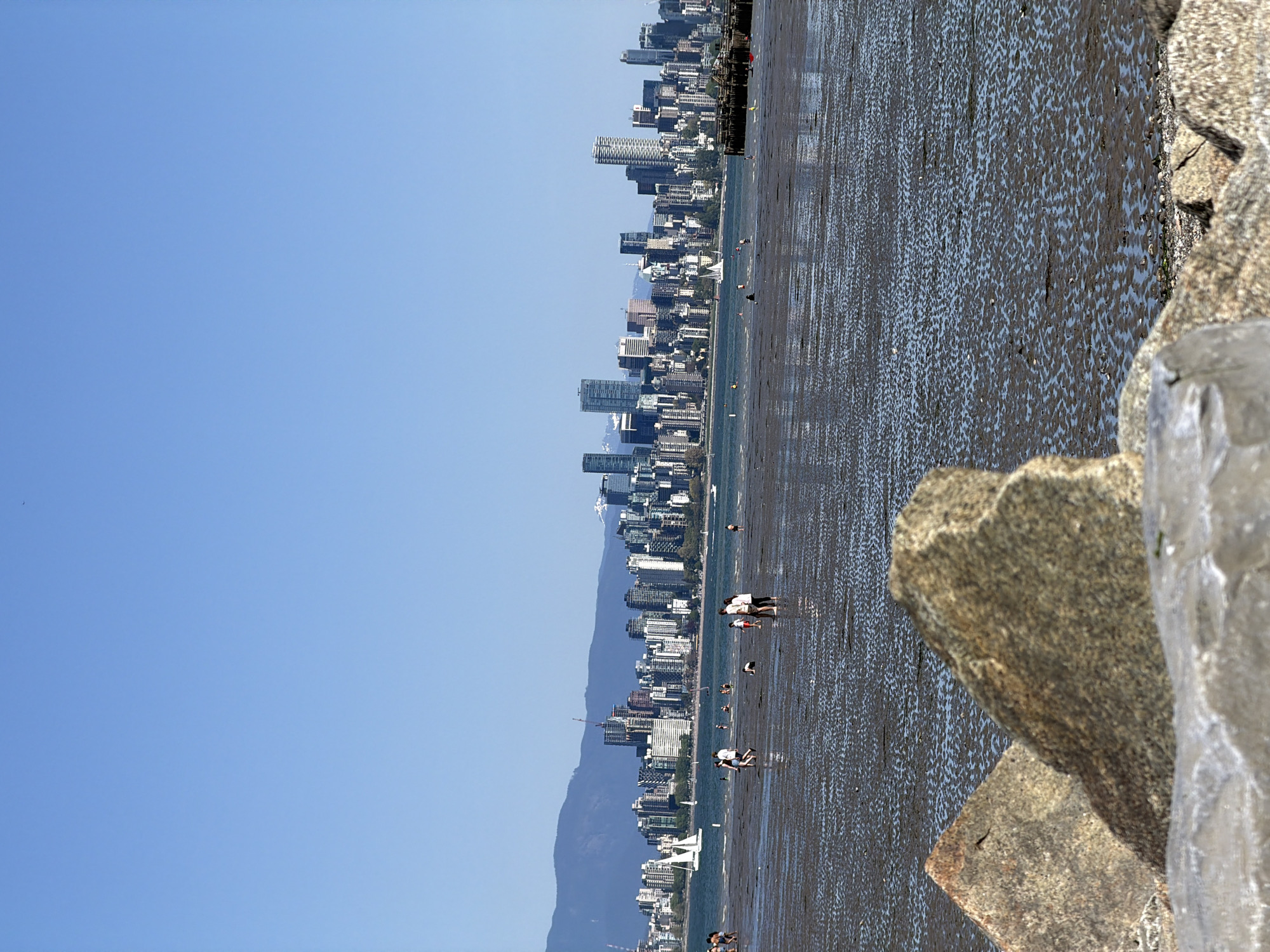 The image captures a view of the Vancouver, Canada skyline from a vantage point on the shoreline. A clear blue sky occupies a significant portion of the left side of the image. The city skyline is visible in the center. The ground is mostly covered with a wet, reflective sand surface. Several small figures of people appear to be walking across the sand. Large rocks are present on the right side of the image.