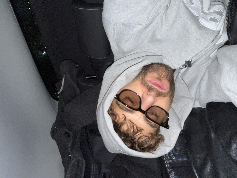 In the city of Givors, France, an adult male is depicted sleeping or resting within the interior of a vehicle. The subject, exhibiting light brown curly hair and a beard, wears a gray zippered hooded sweatshirt with a partially visible white design on the right chest and dark-framed sunglasses with amber-tinted lenses over closed eyelids. His head is positioned against a dark, possibly leather, surface. The vehicle's interior features a light gray roof liner, a dark fabric headrest, and an accumulation of dark fabric items, possibly garments or luggage, on the adjacent seating area. Distant, indistinct lights are visible through a narrow window at the top of the frame, suggesting a nighttime or low-light setting.