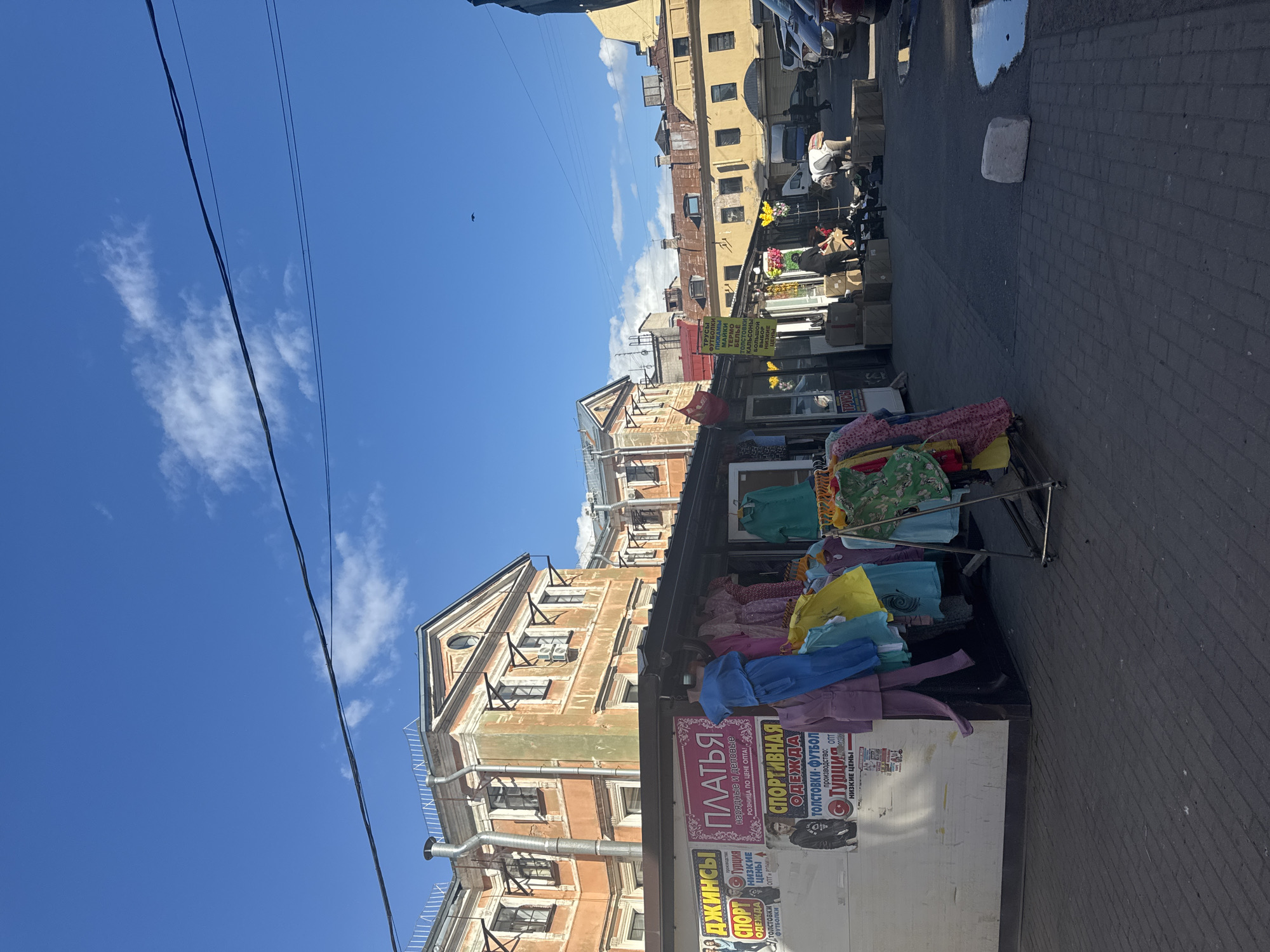 The image, captured in Saint Petersburg, Russia, shows a vibrant street scene with a clear blue sky and scattered white clouds, suggesting a pleasant daytime. The perspective is from a slightly elevated viewpoint, looking down a street lined with old, colorful buildings.
In the foreground, a market stall is prominently displayed, overflowing with a colorful assortment of clothing. Brightly colored shirts, dresses, and pants are neatly arranged on racks and folded on tables. A large white panel on the front of the stall features several advertisements printed in Russian, promoting items like "DRESSES," "SPORTSWEAR," "JEANS," and "TSHIRTS." Some text indicates prices and origin, such as "TURKEY" and "LOW PRICES."
Beyond the market stall, the street continues, revealing more of the urban architecture. The buildings exhibit a mix of architectural styles, with some featuring ornate facades and others appearing more utilitarian. The street itself is paved with cobblestones, and a large puddle reflects the sky, adding to the visual interest of the scene. In the distance, parked cars and a few individuals can be seen, suggesting the hustle and bustle of a market day. The overall impression is one of a lively and bustling street market in a historic Russian city.