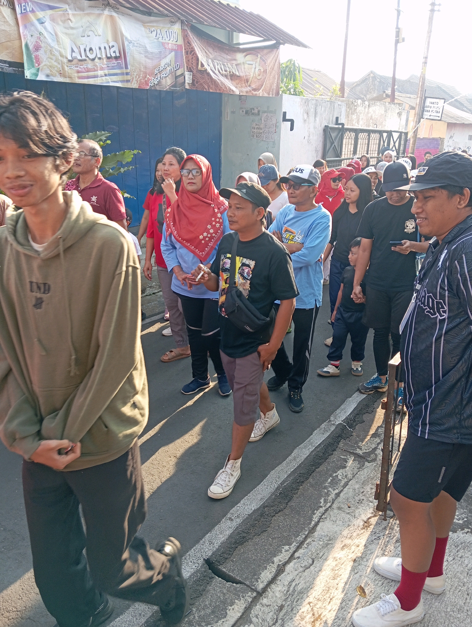 A group of people, predominantly adults and one child, are standing and walking on a street in Magelang, Indonesia. The scene is outdoors, with buildings visible in the background, including a structure with a large banner advertising "Aroma Royal Fusion." The banner also displays a price of "24.000." A smaller sign is also visible on the street with text. The people are dressed casually, with some wearing t-shirts, hoodies, and hats. Some are looking at their phones, while others appear to be interacting with each other. One person in the foreground is holding a cigarette. The ground is asphalt with a white line marking the edge of the road. The lighting suggests it is daytime, possibly late afternoon, as shadows are cast on the ground.