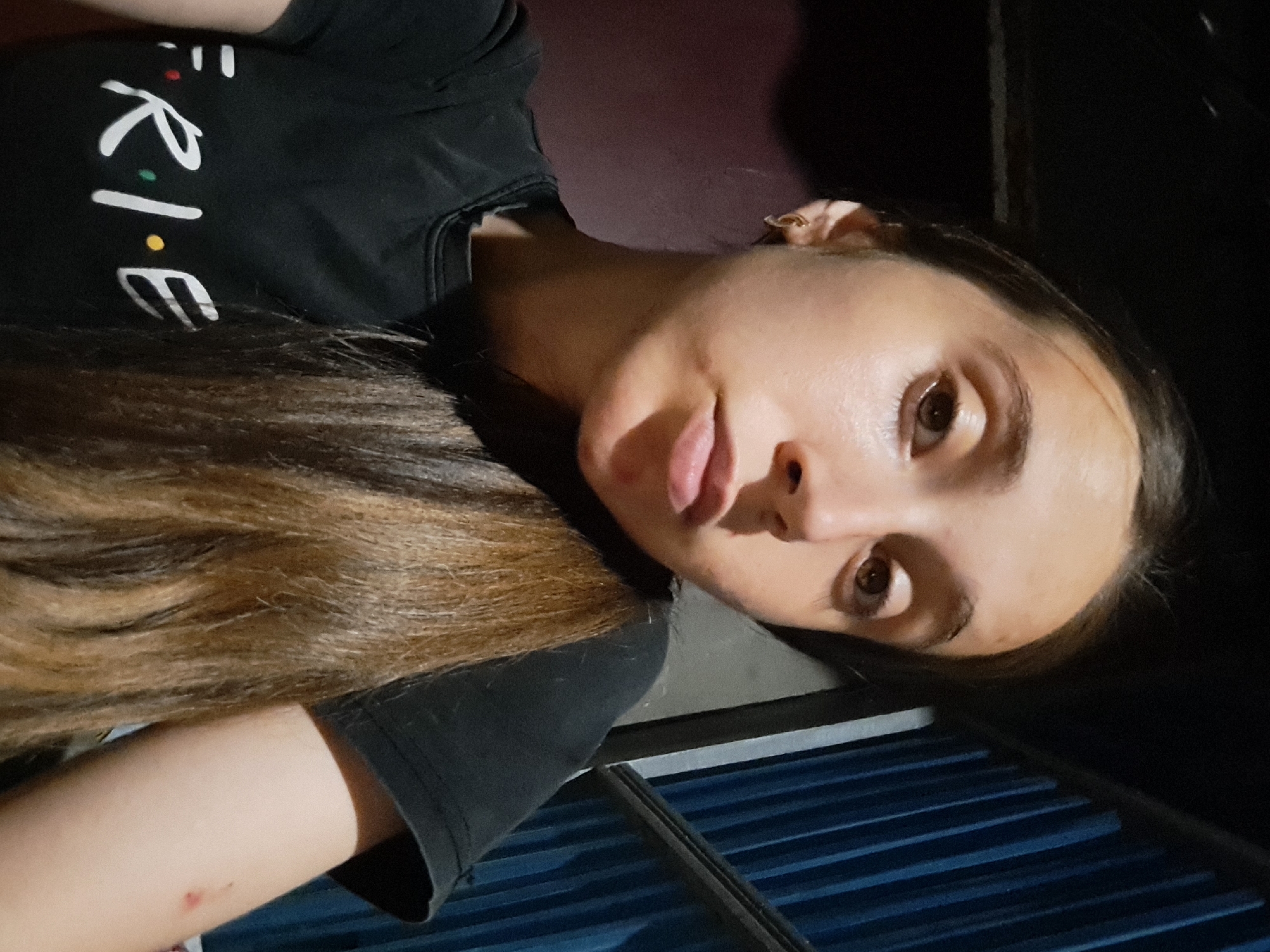 The image is a close-up, selfie-style portrait of a young woman indoors, likely in Vypasne, Ukraine. She has long, light brown hair that falls over her right shoulder. Her skin appears fair, and she is wearing a black t-shirt with the word "FRIENDS" printed in white letters, with colored dots in place of the 'i' and 'e' in the logo. She has a small hoop earring in her left ear. The background is dimly lit and appears to be a room with a dark, possibly wooden wall on the left and a blue, slatted structure on the right, resembling blinds or corrugated metal. The lighting is artificial and casts shadows, suggesting an indoor setting at night or in a poorly lit area. There is a small red mark on her right arm.