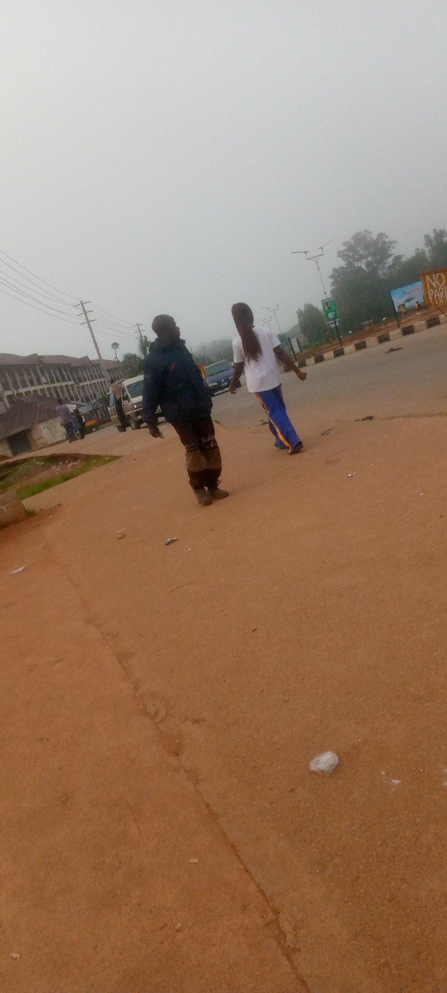 The image depicts a street scene in Gura Top, Nigeria. Two individuals are walking on a dirt path next to a paved road. The person on the left is a male wearing a dark jacket and tan pants. The person on the right is a female wearing a white t-shirt and blue pants with yellow stripes. In the background, there are vehicles, including a van and a car, and buildings lining the street. A "NO PARKING" sign is visible on the right side of the road. The environment appears to be somewhat hazy, suggesting possible fog or low visibility.