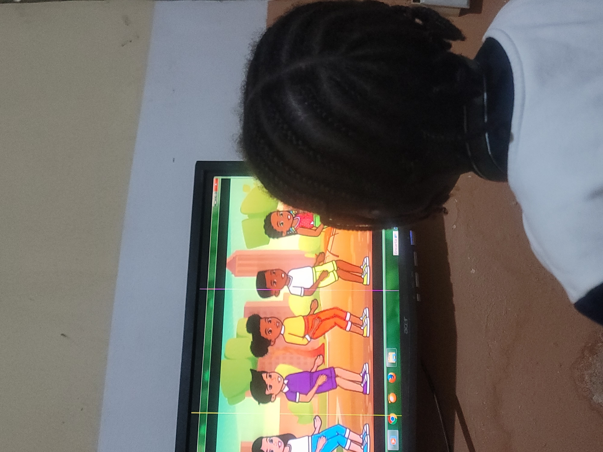 The scene, located in the city of Barakin Akawo, Nigeria, shows a child from behind, facing a computer monitor. The child has dark skin and dark hair styled in braids (cornrows). They are wearing a white long-sleeved shirt with a dark collar and have dark headphones resting around their neck.
The black-bezeled Acer monitor displays an animated scene featuring five characters with diverse appearances, including different skin tones and hair textures, dressed in various colored casual wear (e.g., purple shorts, orange pants, yellow shorts, blue shorts). The characters appear to be in motion or posed dynamically, suggesting a dance or exercise video against a background of green trees and distant city buildings. Vertical yellow and purple lines are visible across the screen. The Windows taskbar is visible at the bottom of the display, showing a play icon, web browser icons (including Chrome, Firefox, Edge, and Opera), and the word "Encarta" in the bottom right corner.
The monitor rests on a light brown, textured surface, possibly a wooden or concrete table. The background features a light grey wall that transitions to a darker, possibly beige, section above the monitor. The wall exhibits minor smudges and blemishes. The floor, partially visible beneath the table, appears to be a similar textured concrete surface.