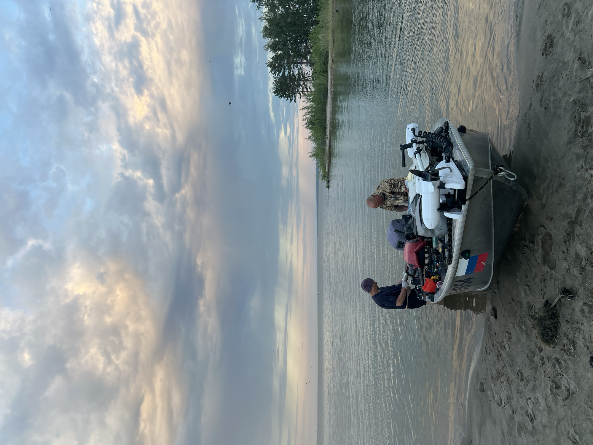 The image depicts an outdoor scene on the shore of a body of water, likely a lake, with a boat partially beached on sandy ground. Two men are present, interacting with the boat and its contents. One man, wearing a dark t-shirt and a head covering, stands to the left of the boat, appearing to be working with something held in his hands. The other man, dressed in a camouflage patterned shirt, is seated in the boat to the right, also engaged with objects within the vessel. The boat is equipped with two electric trolling motors mounted on the stern. A Russian flag emblem is visible on the side of the boat. The environment is characterized by calm water, a cloudy sky with hints of sunset colors, and a distant treeline along the shoreline. The overall impression is one of preparation or activity related to boating or fishing.