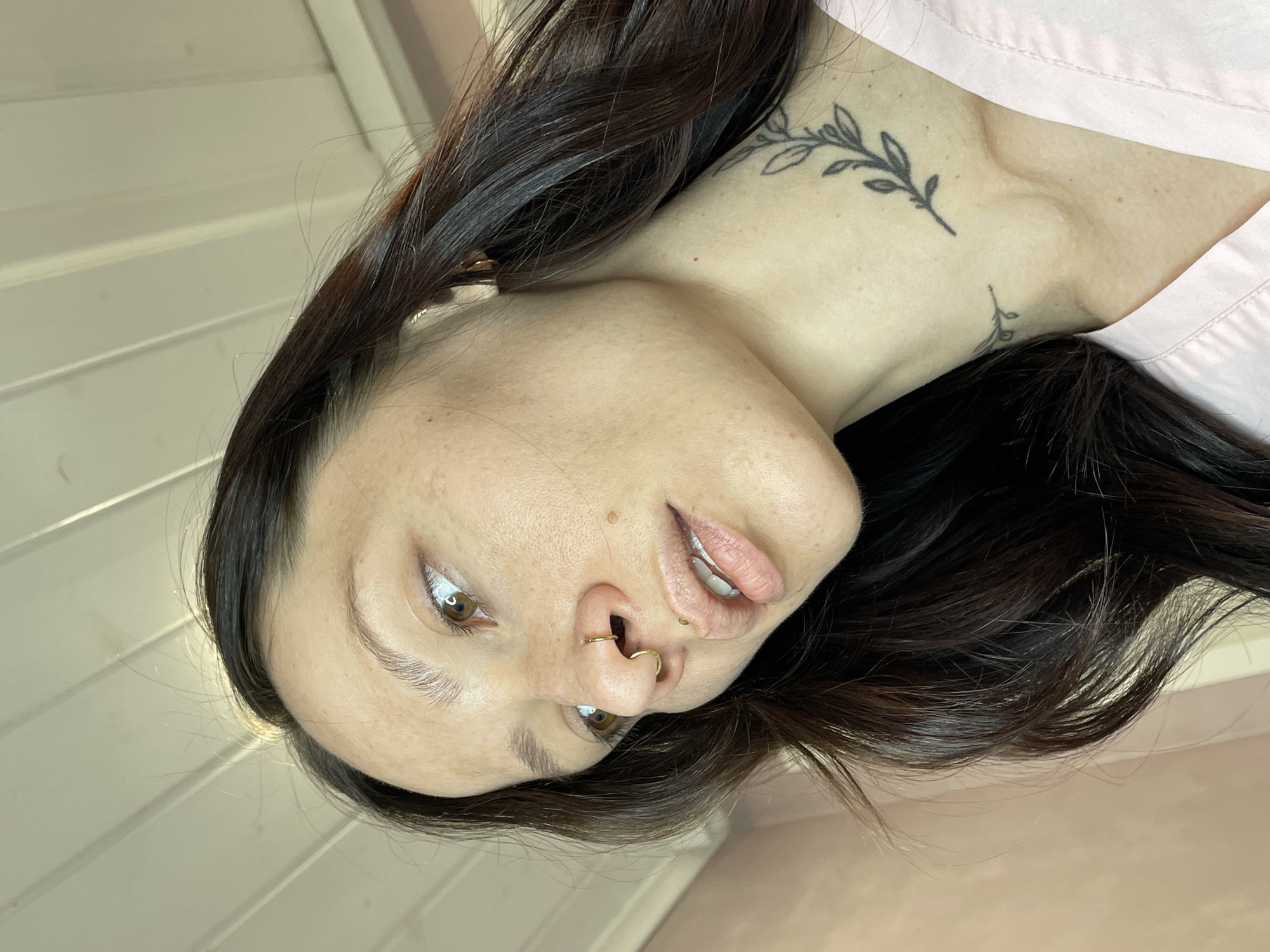 A woman is visible in this close-up, tilted image. She has dark brown hair, light skin, and brown eyes. She wears a gold ring in her nose. A tattoo of a leafy branch is visible on her neck and shoulder. The background includes a glimpse of a white-painted wall and a pink surface. The location is not discernible and cannot be said to be in Izhevsk, Russia.