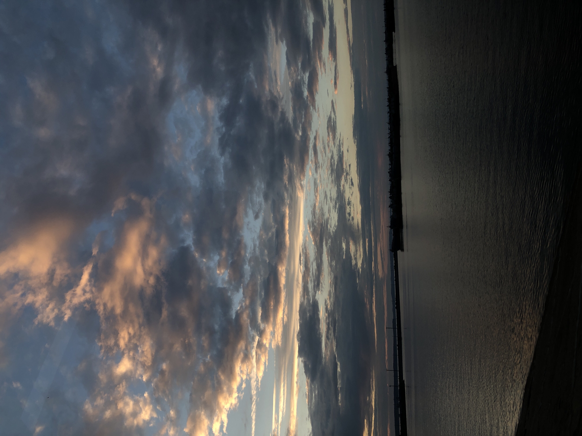 This media file captures a sunset over a body of water, likely a reservoir or large river, with a silhouetted treeline and distant structures along the far shore. The sky is filled with dramatic clouds, a mix of dark, heavy formations and lighter, wispy clouds illuminated by the setting sun. The colors range from deep blues and grays in the shadowed clouds to warm oranges and soft yellows where the light catches them. The water's surface reflects the subdued colors of the sky, appearing dark and calm with gentle ripples. The horizon line is sharp, separating the sky from the water and land. The overall mood is serene and peaceful, characteristic of a sunset. The time of day is clearly evening. There is no visible text in the image. The location context suggests Chaikovsky, Russia, which aligns with the landscape features depicted.