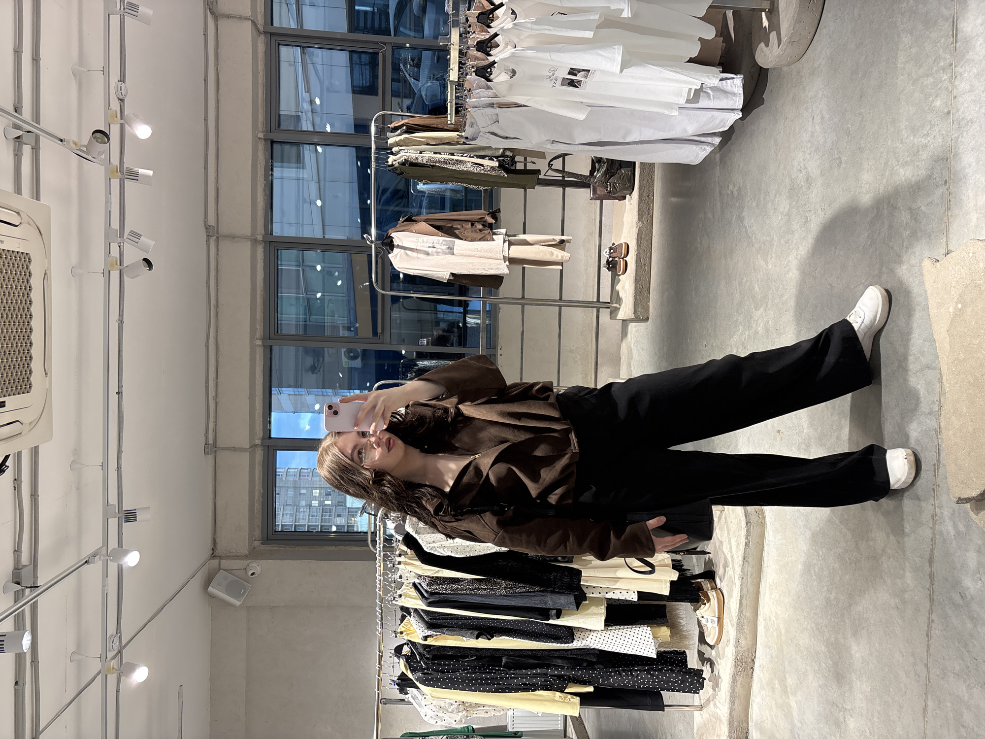 A young woman is pictured inside what appears to be a modern retail clothing store, taking a selfie using her light pink smartphone. She is seen from the chest down, with her face visible in the phone's reflection. She wears a dark brown zippered long-sleeved top, black wide-leg pants, and white sneakers. Her long, dark brown hair falls over her shoulders.
The setting is an indoor space characterized by white walls, a light grey concrete floor, and large windows that offer a view of urban buildings outside. The store features multiple clothing racks. One rack is positioned behind the woman, displaying a variety of garments including t-shirts, blouses, and skirts in neutral tones like white, black, beige, and some with patterns like polka dots. Another rack, partially visible further back, holds more clothes, predominantly in white. Some white t-shirts have indistinct black graphics or text.
The ceiling is fitted with industrial-style track lighting, featuring several white spotlights, some of which are illuminated. An air conditioning unit is mounted high on the left wall. The overall aesthetic is minimalist and modern. The natural light through the windows, combined with the indoor lighting, suggests it could be daytime. The image reflects an ordinary activity of a customer in a fashion store, likely in Perm, Russia.