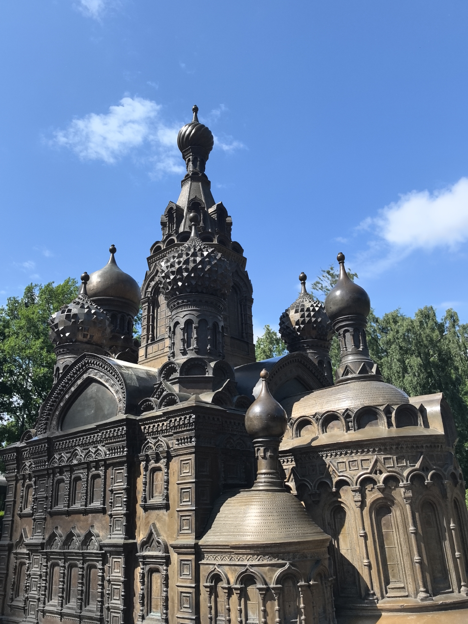 This is a medium shot, eye-level photograph of a miniature bronze replica of a Russian Orthodox church, likely a representation of a famous landmark in Saint Petersburg, Russia. The sculpture is intricately detailed, showcasing multiple domes of varying sizes, adorned with ornate patterns and finials. The facade features rows of small windows and decorative architectural elements, suggesting a historical and religious building. The bronze material gives the sculpture a dark, weathered appearance, reflecting sunlight and creating shadows that highlight its detailed craftsmanship.
The church model is positioned outdoors, with a clear blue sky and scattered white clouds serving as the backdrop. Lush green trees are visible behind and to the sides of the sculpture, suggesting a park or a garden setting, common for such public art installations. The bright sunlight indicates it is daytime, likely midday given the strong illumination and clear skies. No people are visible in the frame, placing the focus solely on the architectural model and its surroundings.
The detailed design of the church, with its multiple onion domes and intricate facade work, is characteristic of traditional Russian church architecture. The setting in Saint Petersburg is a plausible context for such a replica, given the city's rich history and numerous iconic religious structures. The overall mood is one of serene contemplation, highlighting the beauty and historical significance of Russian religious architecture, presented in a miniature, artistic form. There is no visible text in the image.
