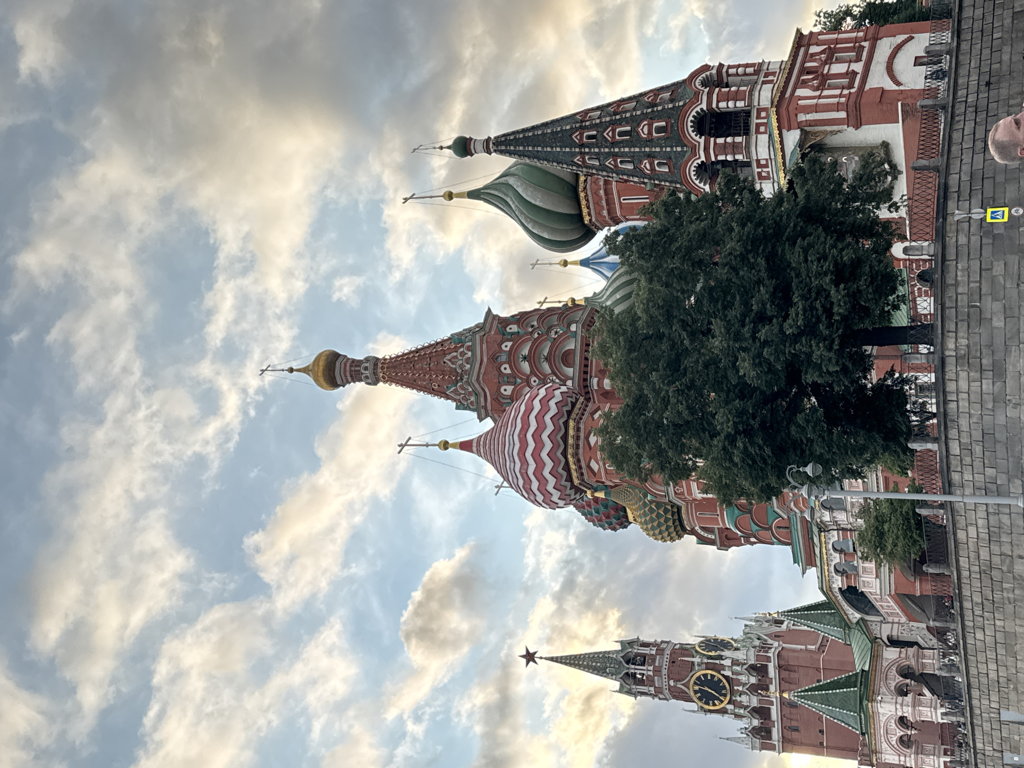 The image captures a low-angle view of Saint Basil's Cathedral and the Spasskaya Tower in Moscow, Russia, under a cloudy sky with shafts of sunlight breaking through. The iconic onion domes of Saint Basil's Cathedral, painted in vibrant red, white, and green patterns, are prominently featured, along with intricate architectural details. To the left, the Spasskaya Tower, a red brick clock tower topped with a star, stands tall. A large tree with dense foliage is positioned in the foreground, partially obscuring the cathedral. A stone wall with a metal railing runs along the bottom of the frame, and a street lamp is visible on the right. A man's head and shoulders are visible at the bottom right corner of the image, looking towards the right. The overall scene depicts a notable landmark in Moscow during what appears to be late afternoon or early evening.