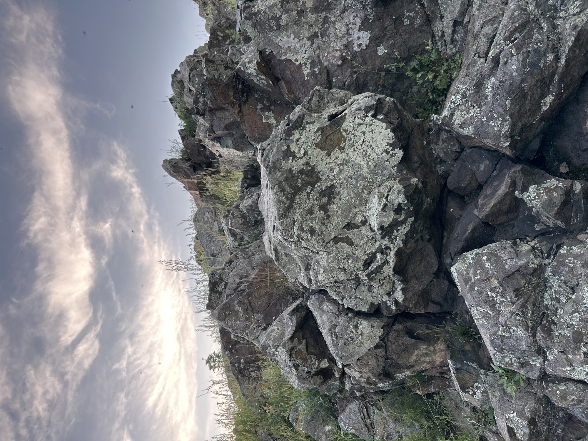 This is a low-angle, close-up, outdoor photograph of a rocky outcrop. The foreground is dominated by large, weathered rocks, some covered in patches of light-colored lichen. Sparse dry grass and small green plants grow between and around the rocks. In the background, the sky is visible, filled with wispy, light-colored clouds against a pale blue expanse. The lighting suggests it may be either dawn or dusk. There are no people or animals visible in the image. The location is described as Unknown, Unknown.