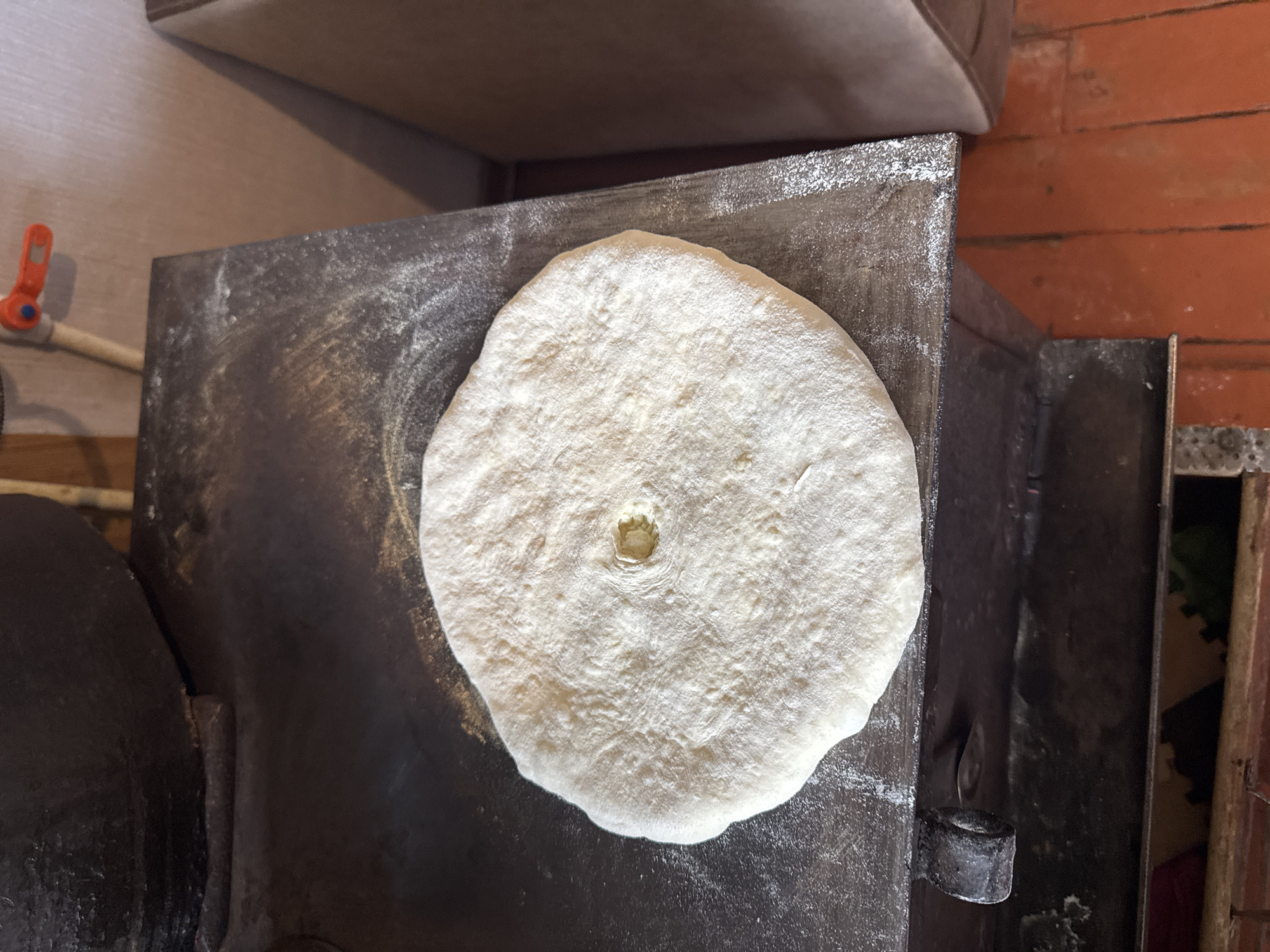 A flatbread, dusted with flour, rests on a dark, worn metal surface. The surface appears to be the top of a wood-burning stove, as evidenced by the black, sooty material and a metal handle on the left side. The flatbread has a small, indented circle in its center. The background shows a textured, light-colored wall and a portion of a brown upholstered object, possibly a chair or sofa. The floor below is made of reddish-orange tiles. The scene is likely in a kitchen or cooking area. The image does not provide enough information to determine the exact location in Tandzia, Georgia.