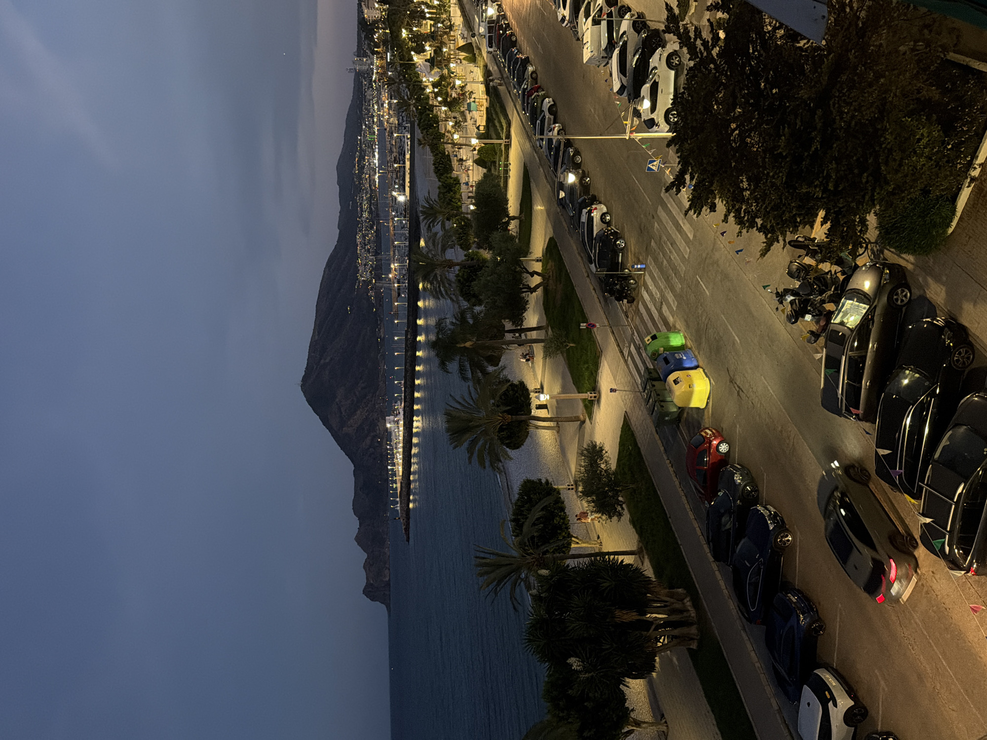 The image captures an evening scene in Altea, Spain, looking out over the Mediterranean Sea towards the coastline. The sky is a deep twilight blue, with a few faint clouds. In the foreground, a wide street lined with parked cars runs parallel to the coast. The cars are a mix of colors and models, with headlights and taillights illuminated in some cases. Across the street, a beach is visible, with palm trees and streetlights casting a warm glow. Further out to sea, a pier with lights stretching along its length juts out into the water, with a distant hillside town illuminated by numerous lights. The overall impression is of a coastal town at dusk, with the gentle lights of civilization contrasting with the dark expanse of the sea and sky.