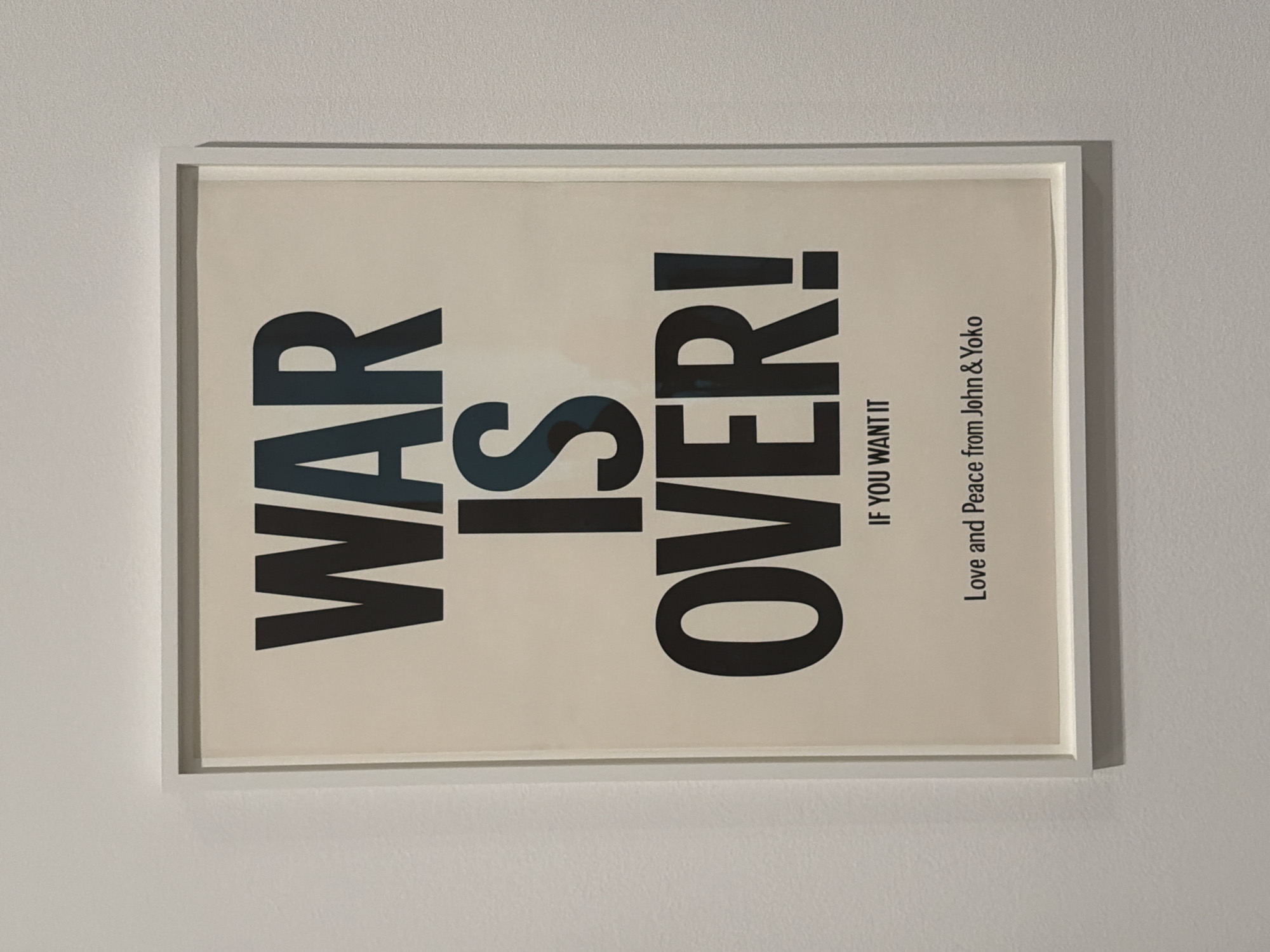 The image shows a framed print of the iconic "War Is Over! If You Want It" poster by John Lennon and Yoko Ono. The poster features large, bold black lettering against a light background. Below the main text is the smaller inscription "Love and Peace from John & Yoko." The poster is in a white frame and mounted on a pale gray wall. The image provides no information to determine location in Los Angeles, United States of America.
