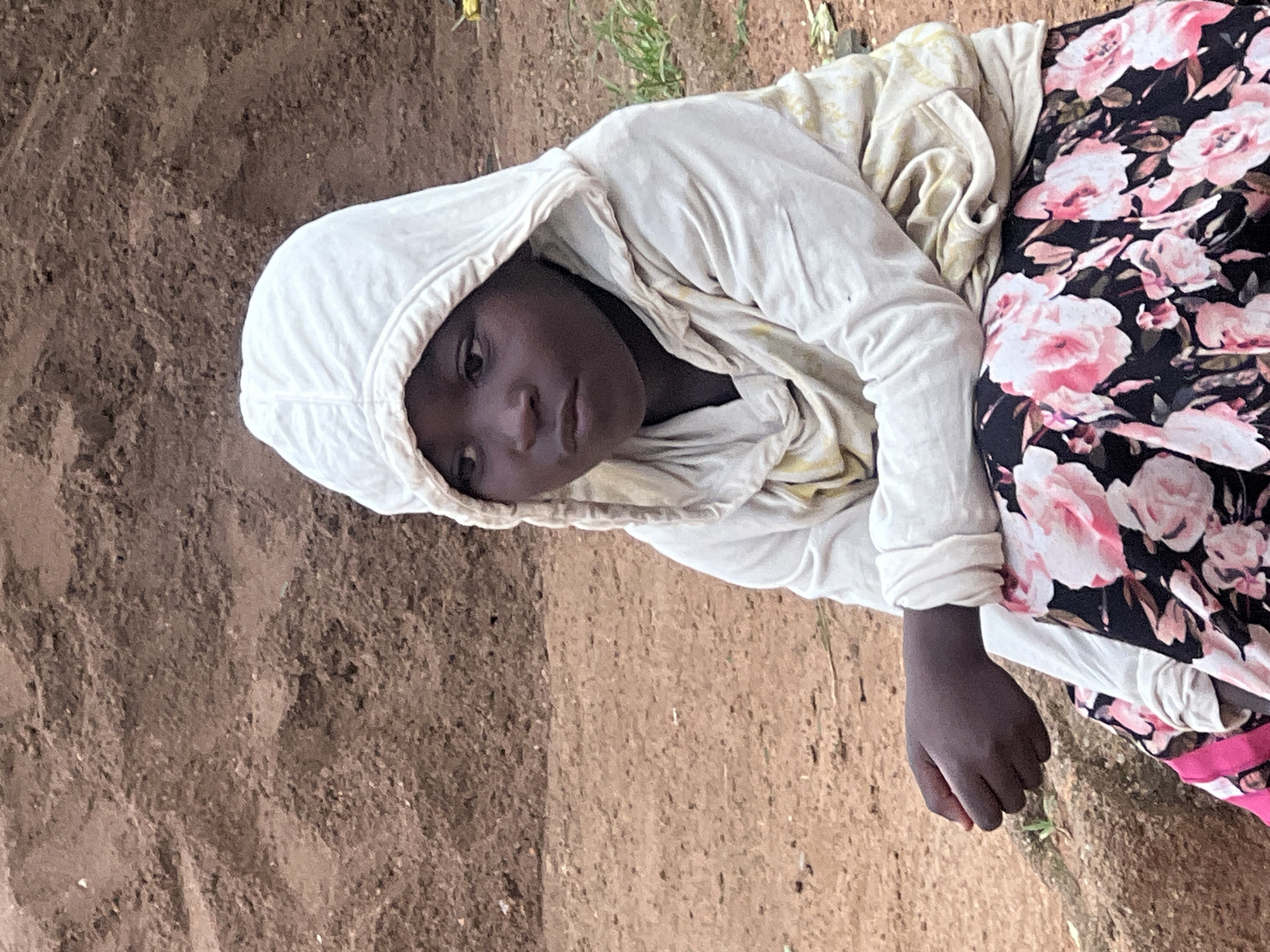 A young Black child, likely a girl given the floral dress, is shown from a slightly elevated angle. The child is wearing a white hooded garment with a yellow patterned undershirt, and a dark fabric dress with a prominent pink and white floral pattern. The child's dark skin is visible, and their left hand, with closed fingers, rests on the ground. The child's face is turned towards the camera, and they appear to be looking directly at it with a neutral or perhaps slightly solemn expression. The hooded garment obscures much of the child's hair and forehead.
The child is positioned on a surface that appears to be dry, brown earth, possibly a dirt path or bare ground, with some sparse, dry vegetation visible in the background. The lighting suggests it is daytime, with natural light illuminating the scene. The overall impression is of a candid moment captured outdoors. The backdrop is predominantly brown earth, suggesting a rural or semi-rural setting in Jos, Nigeria. There is no visible text in the image.