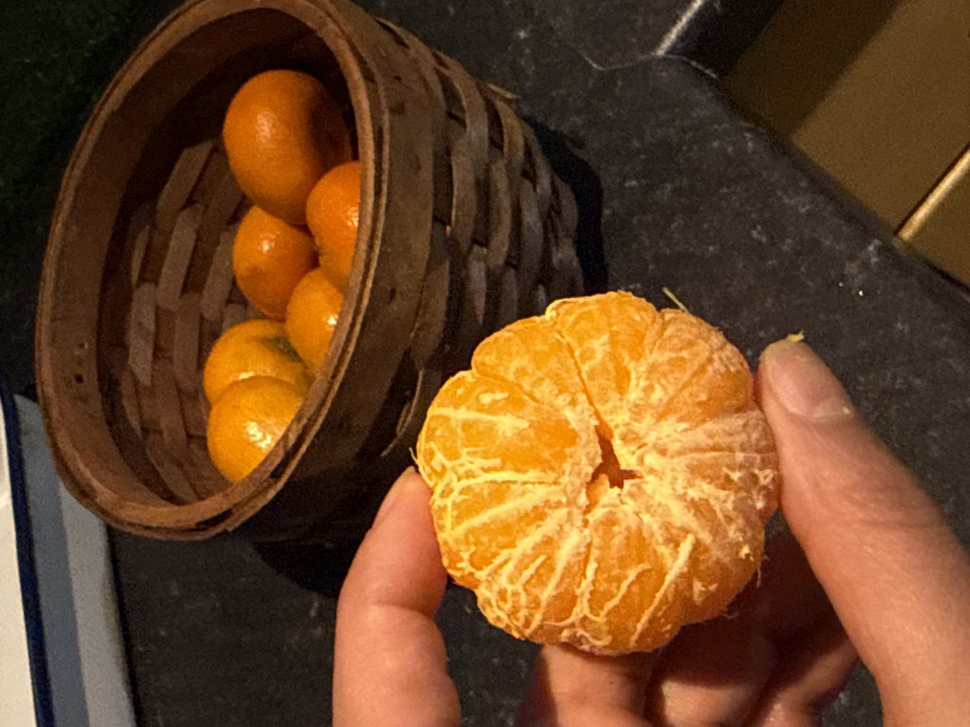 A close-up, angled shot depicts a hand holding a peeled tangerine, positioned next to a small, woven basket filled with more unpeeled tangerines. The backdrop is a mottled, dark grey surface. The image does not contain sufficient information to definitively determine the location as Great Yarmouth, United Kingdom, nor does it depict people, actions, or interactions beyond the basic holding of the fruit.