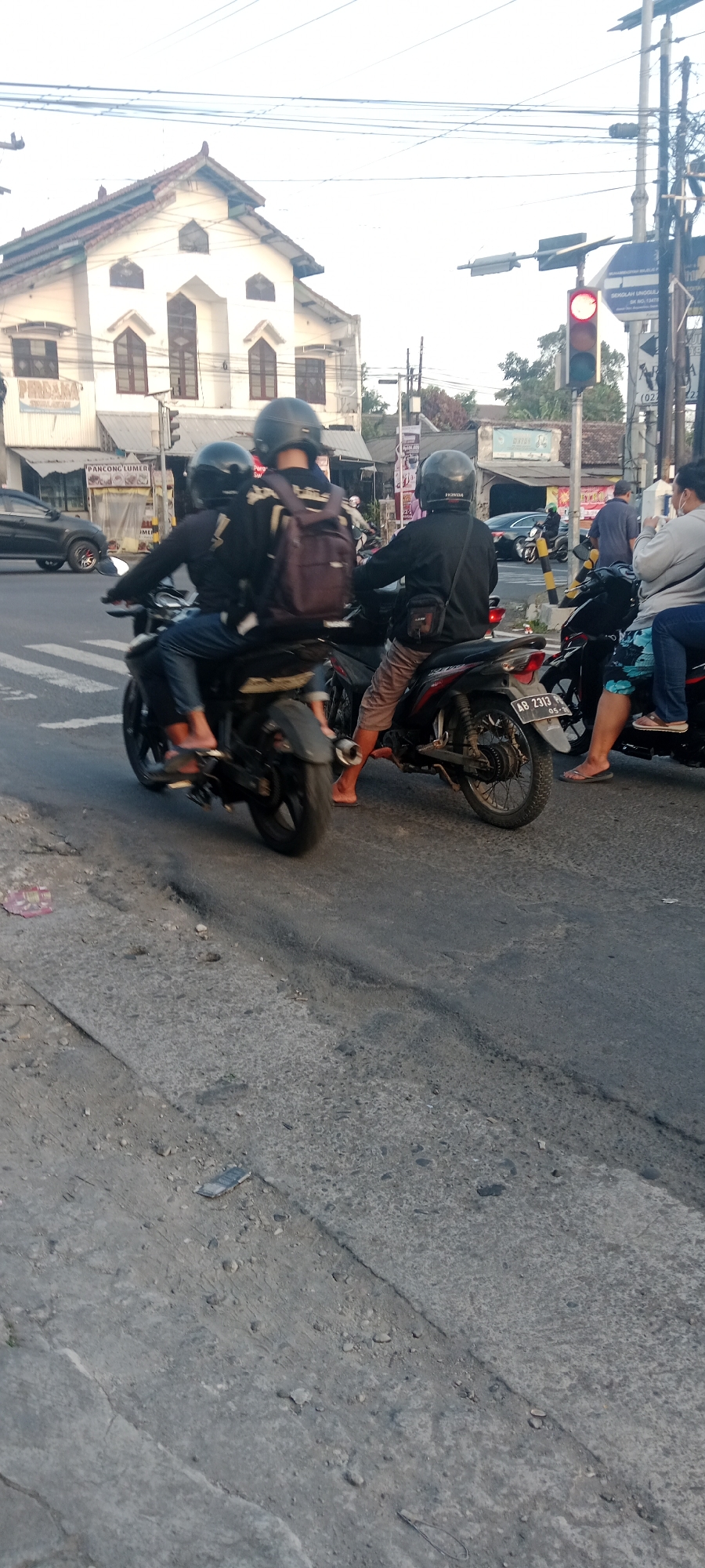 In the city of Maguwoharjo, Indonesia, a street scene unfolds at an intersection with a traffic light. The sky is bright, indicating daytime. In the foreground, the asphalt road shows signs of wear with cracks and debris. A black car is visible in the background on the left.
The primary focus of the image is on several individuals riding motorcycles. On the left, a motorcycle carries three people, all wearing helmets. The driver and the passenger behind them are wearing black long-sleeved shirts, while the person in the very back is also wearing a helmet and a backpack. They are positioned on a crosswalk.
To the right, another motorcycle is occupied by two people, both wearing helmets. The driver is wearing a black long-sleeved shirt and brown shorts, with a small bag slung across their chest. The passenger behind them is wearing a light-colored shirt and appears to be holding something.
Further to the right, a woman wearing a face mask and a gray top is seated on a motorcycle. She is looking away from the camera.
A red traffic light is illuminated, suggesting a halt in traffic. Buildings line the street in the background, including a white building with arched windows. Other structures and street furniture, such as traffic signs and utility poles, are also present. The overall impression is a snapshot of daily life at a busy intersection in an Indonesian city.
