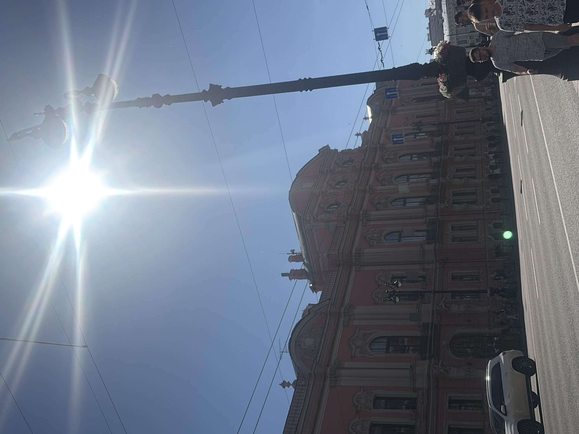 The image shows a street scene in Saint Petersburg, Russia, under a bright, sunny sky. The sun is very strong, creating lens flares that obscure parts of the image.
The main subjects are a building with ornate architecture, a street with a traffic light, and several people. The building appears to be historic, with detailed facades and arched windows. A lamppost stands in front of it. On the street, a white car is parked. Several people are walking on the sidewalk and across the street.
The activity appears to be a normal day with people going about their business. The bright sun suggests it is daytime, likely midday or afternoon. The presence of a traffic light and the paved street indicate an urban environment. The distinctive architecture points to the specific location in Saint Petersburg.
There is no visible text in the image. The overall mood is bright and energetic, due to the strong sunlight and the activity of the people.