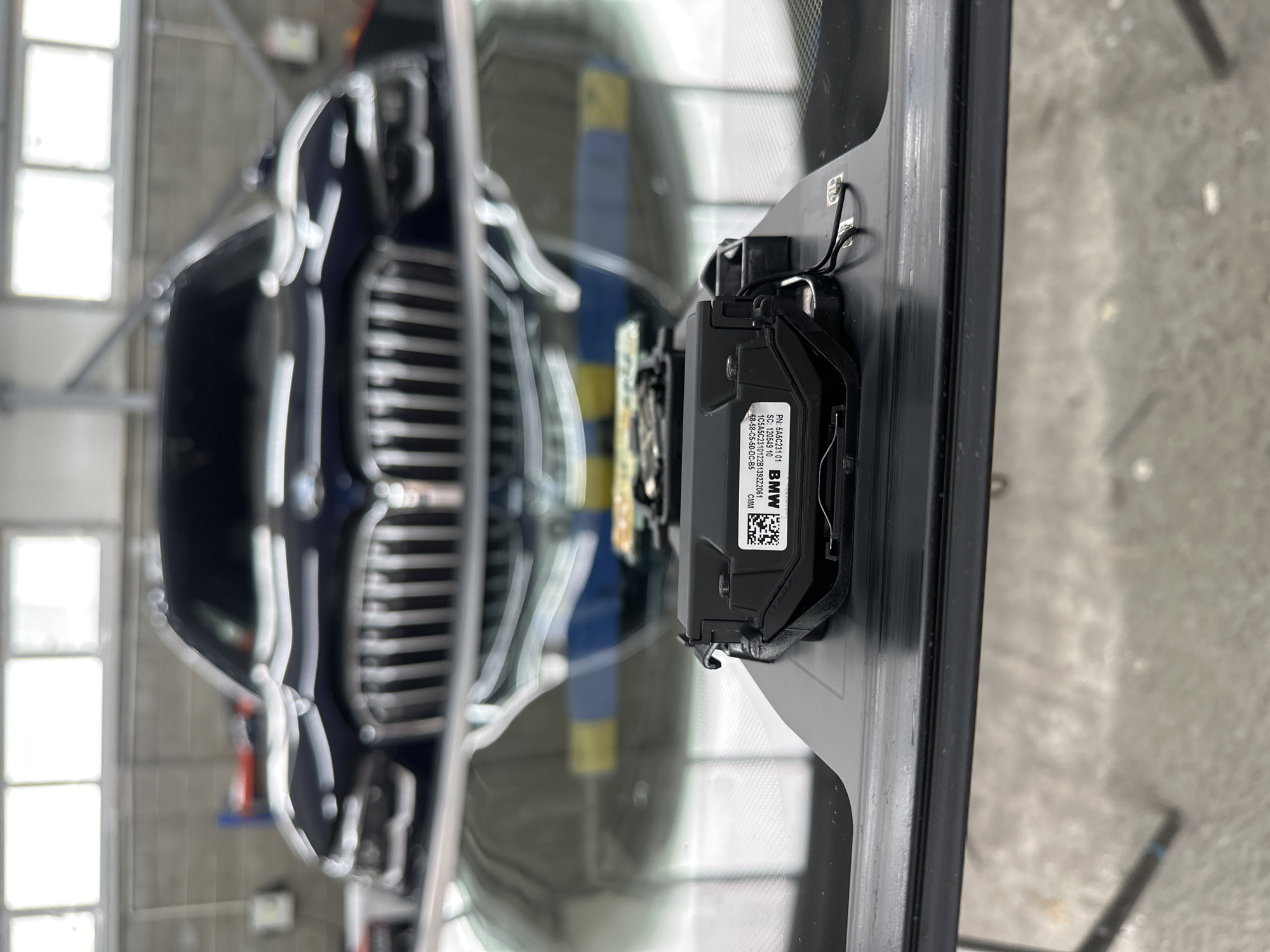 The image shows a close-up of a black electronic device mounted on the interior of a car's windshield. The device has a label with text and a QR code, and wires are attached to it. In the background, the front grille and headlights of a dark-colored BMW SUV are visible through the windshield, with the car parked indoors. The overall environment appears to be a garage or service bay, suggested by the industrial lighting and concrete floor.