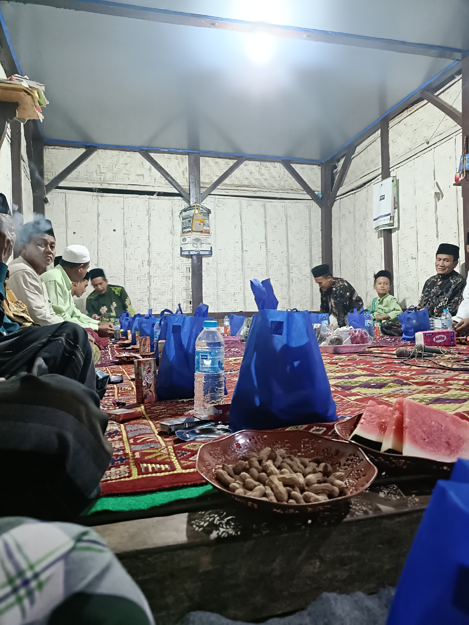 The image depicts an indoor gathering of men and boys, likely a religious or social event, in Bicorong, Indonesia. They are seated on the floor on a patterned rug, with several blue shopping bags and a bottle of water placed on it. In the foreground, a bowl of peanuts and slices of watermelon are visible. The men are dressed in traditional Islamic attire, including peci caps. One man in the left foreground is smoking a cigarette. The room appears to be a simple structure with a wooden frame and woven walls. The lighting suggests it is nighttime or a dimly lit indoor space.
