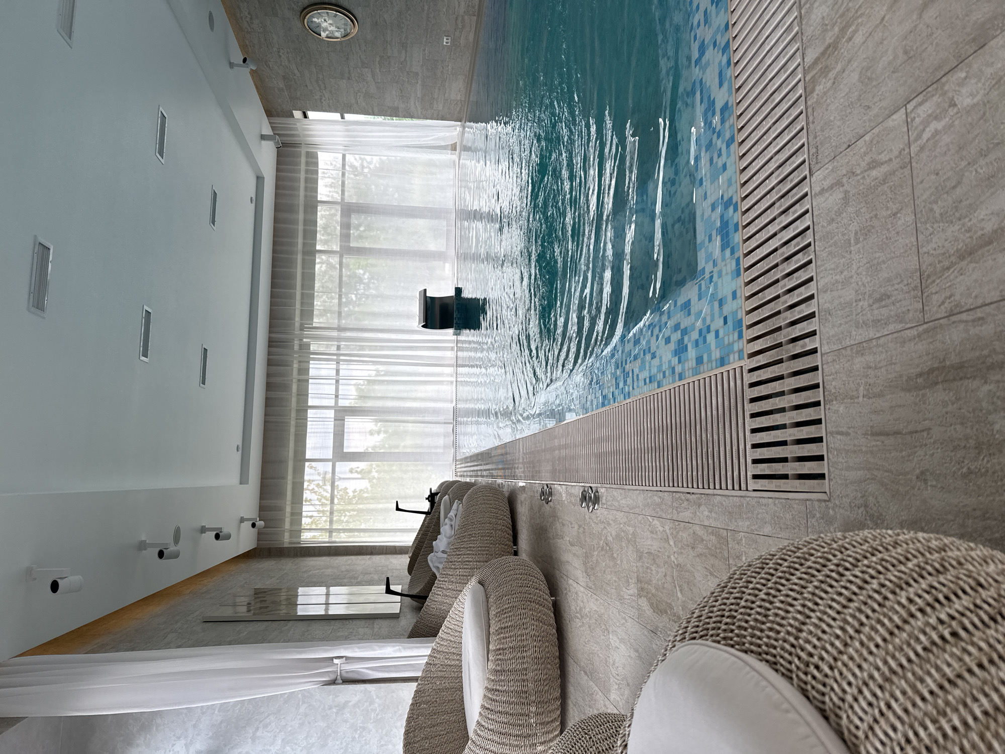 This is a photograph of an indoor swimming pool with a modern design. The pool features a tiled edge with a metallic grate along the perimeter. The water is a vibrant blue, and ripples indicate movement.
In the foreground, to the left, are several light-colored woven chairs with cushions. White sheer curtains hang from the wall behind them, covering large windows that offer a view of greenery outside. The walls of the pool area are textured, with some sections appearing to be stone or concrete.
The ceiling is white and features recessed lighting and air vents. A clock with a gold frame is visible on the wall in the upper right portion of the image. A dark object, possibly a fountain or decorative element, is situated in the pool.
The overall impression is one of a clean, relaxing, and well-maintained indoor swimming facility. The location is identified as the city of Razumnoye, Russia.