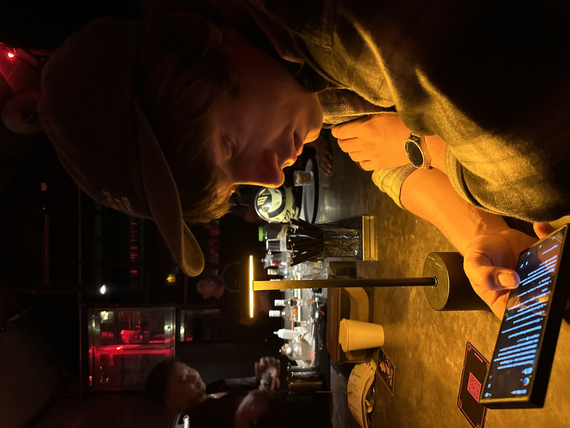 A person wearing a denim jacket and a baseball cap is sitting at a bar in Canggu, Indonesia, looking at a smartphone. The bar has a glass display case with red lighting and a variety of bottles on the shelves behind the bar. The person is wearing a watch on their left wrist, and there is a black stand-up lamp on the counter beside them. A stack of papers, a white cup, and a card with a QR code are on the counter next to the phone. Other people are also at the bar, visible in the background.