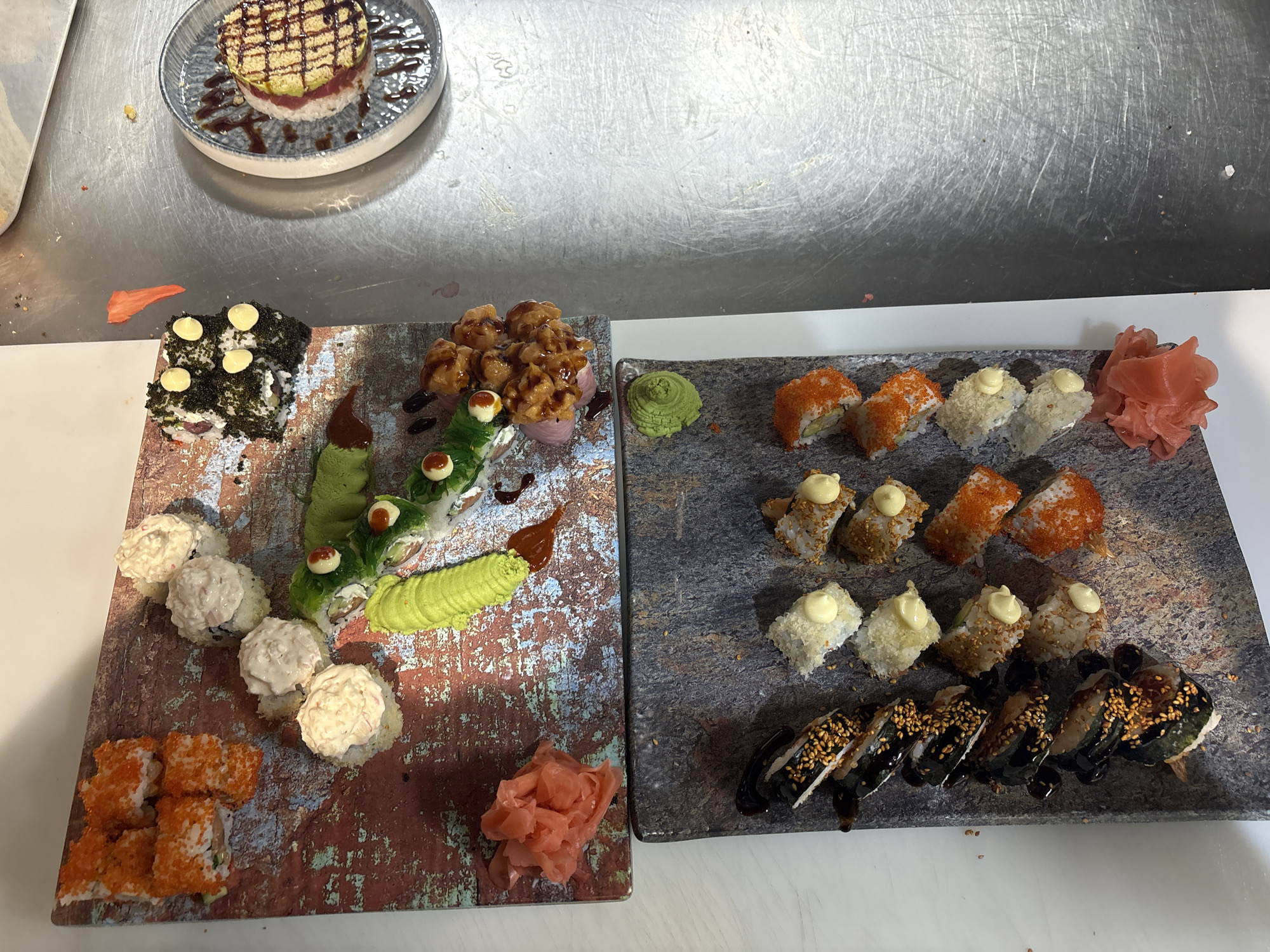 The image shows a tabletop display of sushi and Japanese cuisine. On the left, a square stone slab features an assortment of sushi rolls, including those topped with fish eggs, creamy sauces, and a roll resembling a caterpillar, along with dollops of wasabi and pickled ginger. Another stone slab to the right presents additional varieties of sushi, including those with orange roe, creamy toppings, and eel drizzled with sauce and sesame seeds, alongside a portion of wasabi and pickled ginger. At the top left of the image, a plate contains a stacked appetizer consisting of what appears to be tuna, rice, and avocado, drizzled with sauce. The background appears to be a stainless steel counter or workspace. The photograph was taken in an unidentified location in the city of Burgas, Bulgaria.